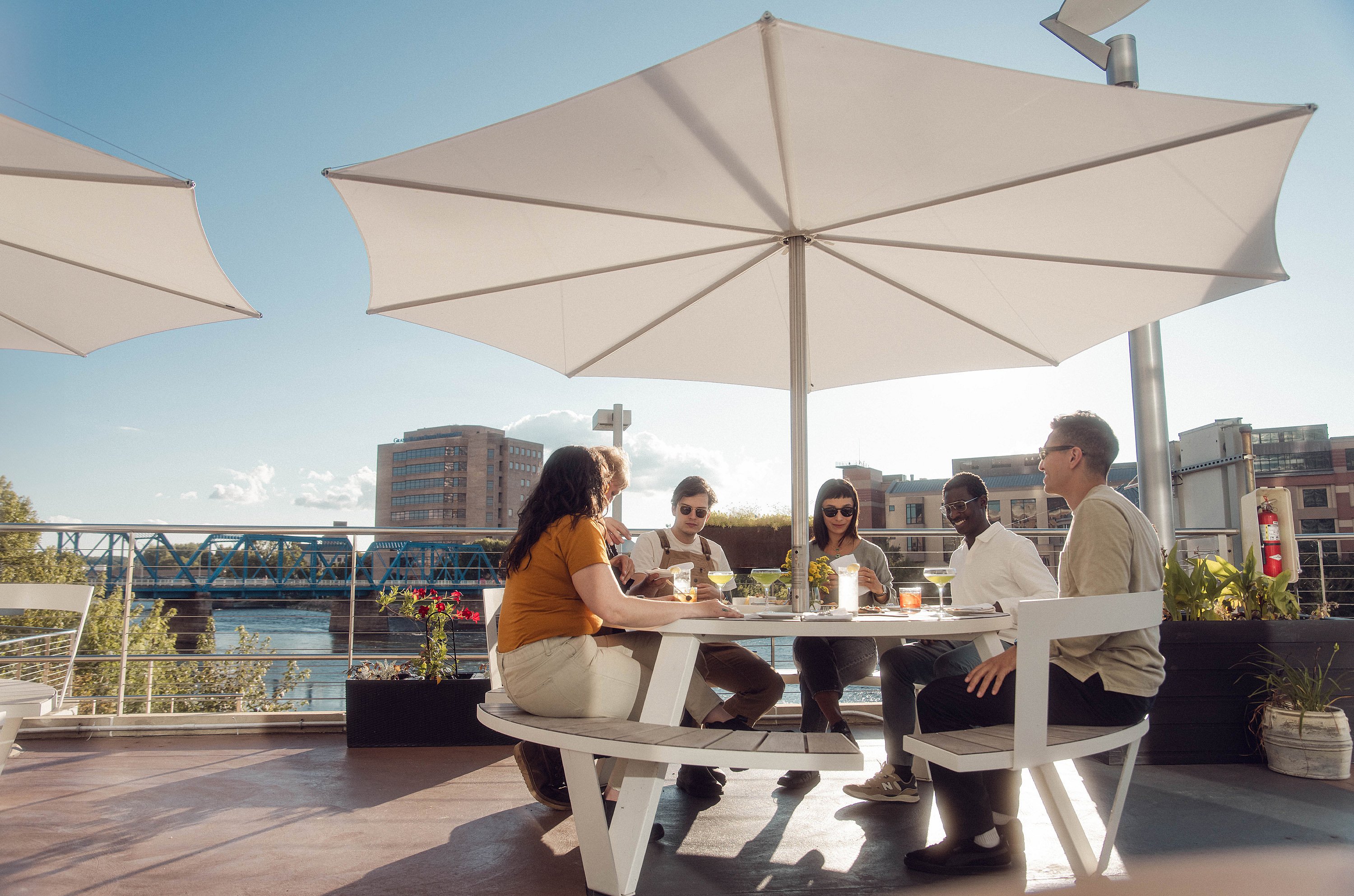 Cinq amis se sont rassemblés autour d’une table blanche de Pantagruel sous une inumbra blanche, partageant des boissons sur le toit-terrasse avec la ligne d’horizon de la ville derrière eux.