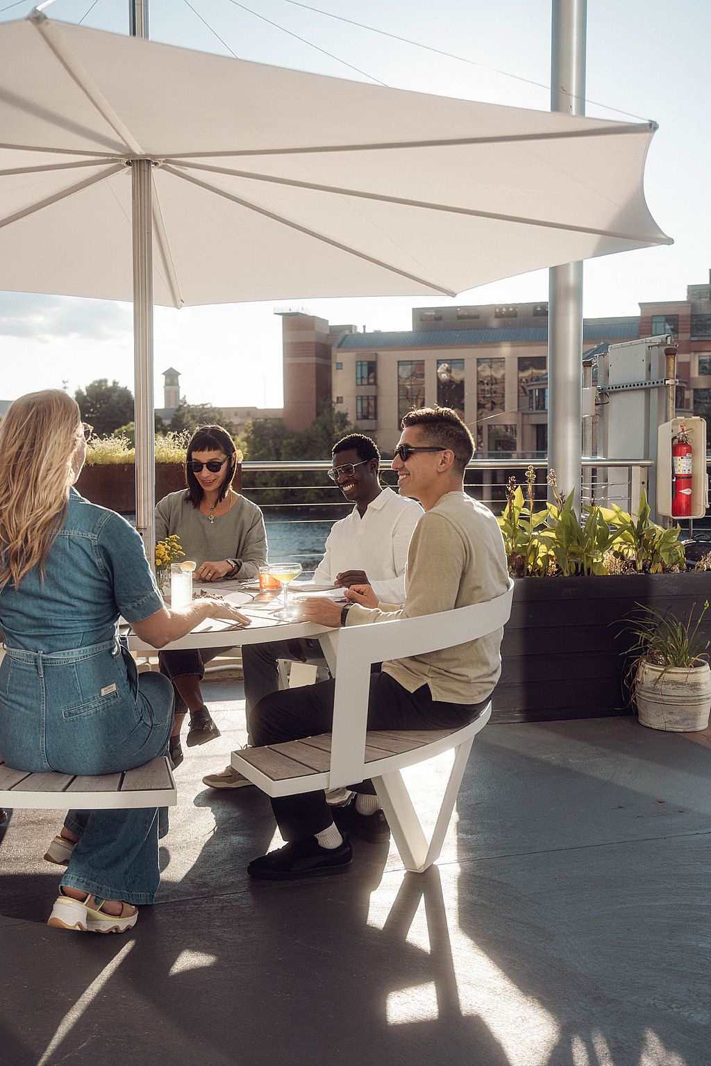 Quatre invités dînent à une table ronde blanche en plein air sous un grand parasol blanc, avec des jardinières et le paysage urbain au bord de la rivière derrière eux.