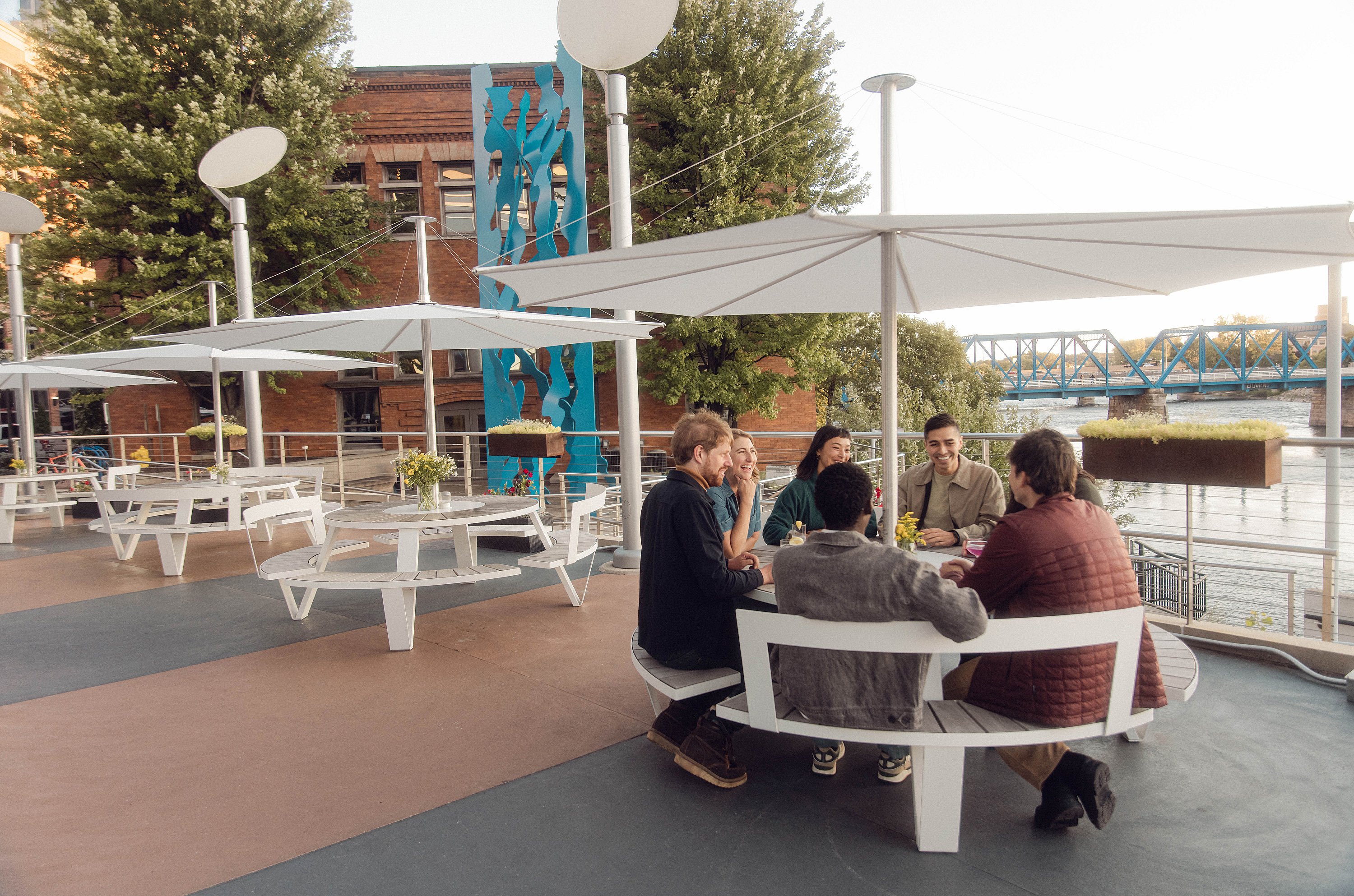 Vriendengroep zitten rond een witte ronde Pantagruel picknicktafel onder een grote witte Inumbra parasol op het JW Marriott dakterras, met nog een bijpassende tafel en stadsgebouwen op de achtergrond.