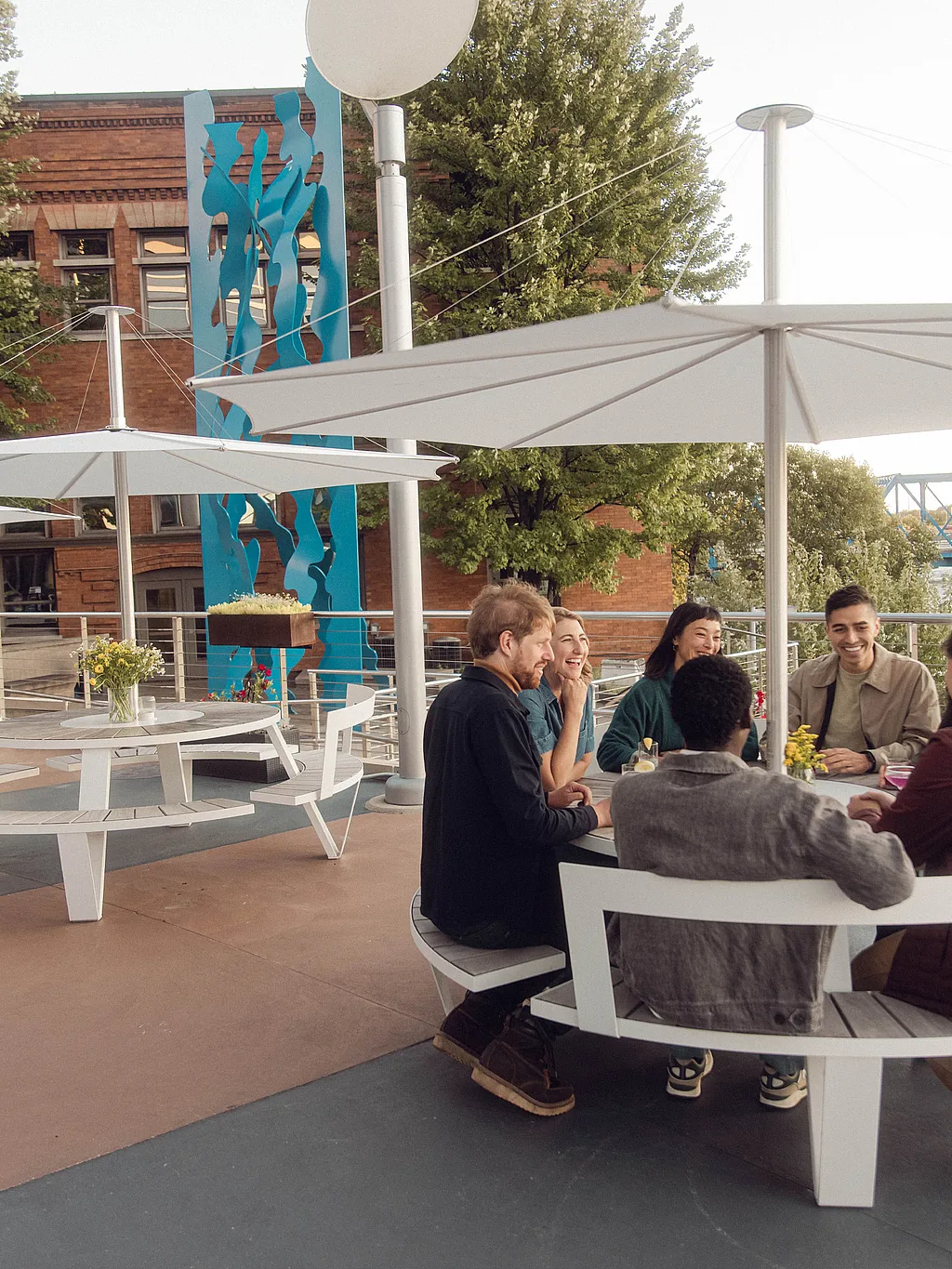 Vriendengroep zitten rond een witte ronde Pantagruel picknicktafel onder een grote witte Inumbra parasol op het JW Marriott dakterras, met nog een bijpassende tafel en stadsgebouwen op de achtergrond.
