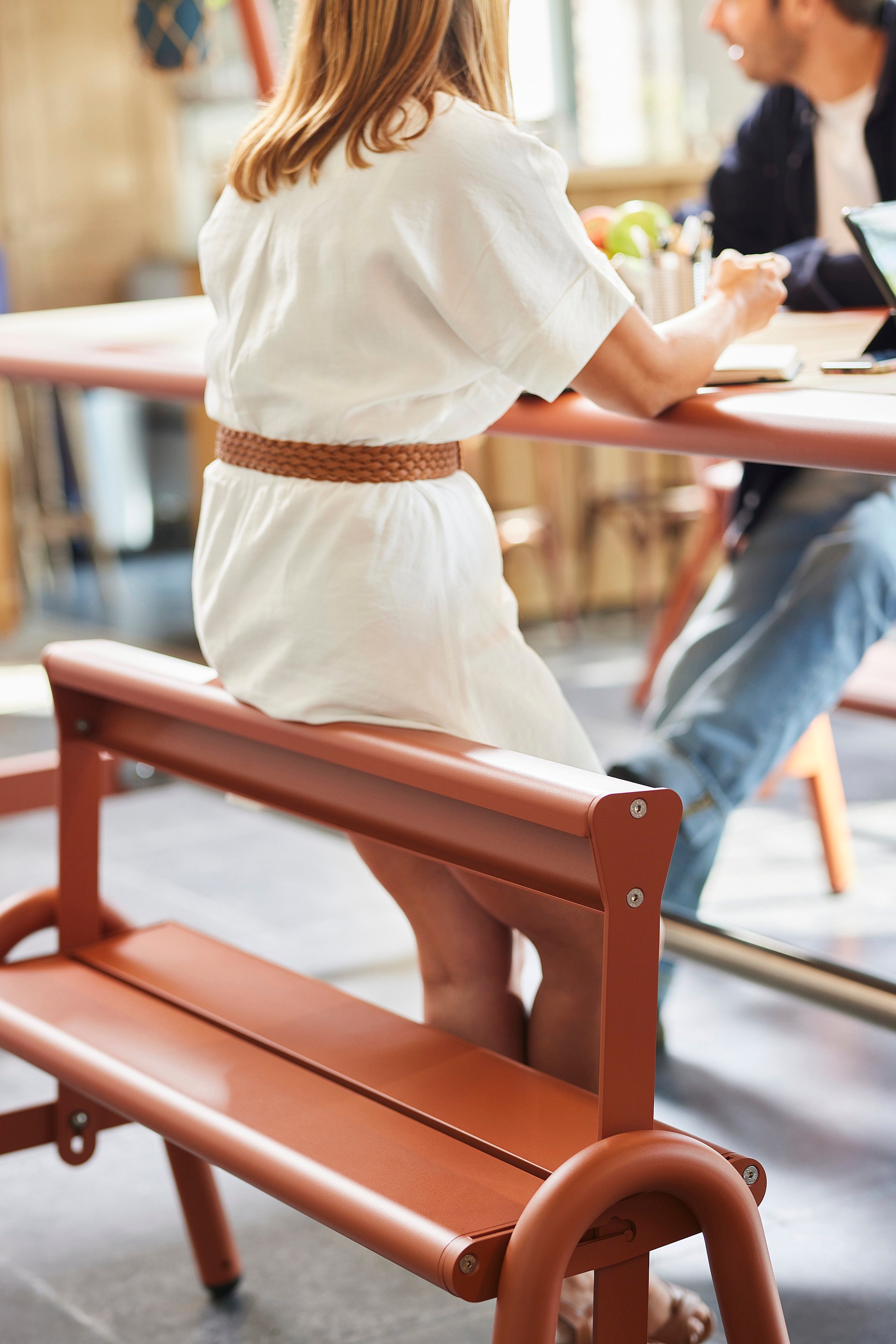 Vrouw in een witte jurk zittend op een oranje bankje aan de werktafel, met de rugleuning en stoelconfiguratie terwijl collega's tegenover haar samenwerken.