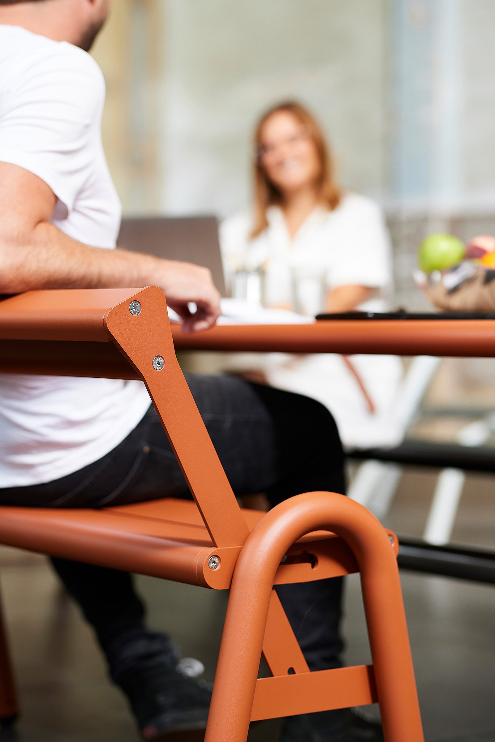 Close-up van een persoon die op een oranje verstelbare bank zit met rugleuning aan de coworking-tafel, met een andere collega op de achtergrond wazig.