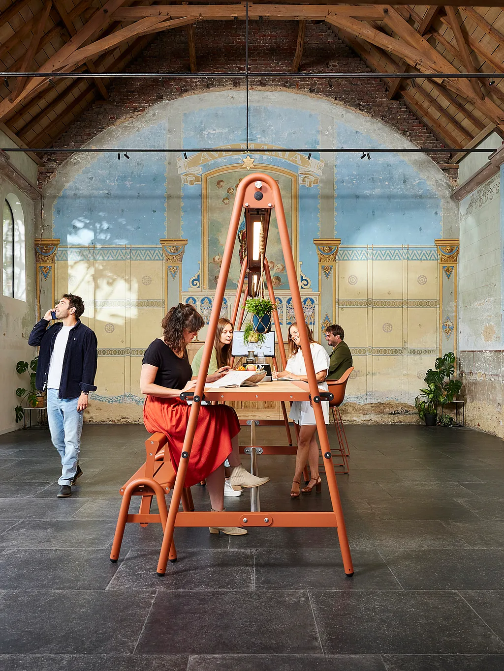 Kollegen verwenden einen orangefarbenen AMAi-Tisch in einer restaurierten Kapelle mit hohen Holzsparren, Bogenfenstern und einer verblassten blauen Wandwand dahinter.