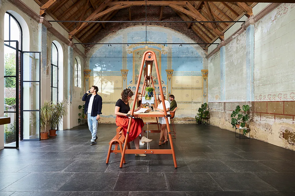 Kollegen verwenden einen orangefarbenen AMAi-Tisch in einer restaurierten Kapelle mit hohen Holzsparren, Bogenfenstern und einer verblassten blauen Wandwand dahinter.