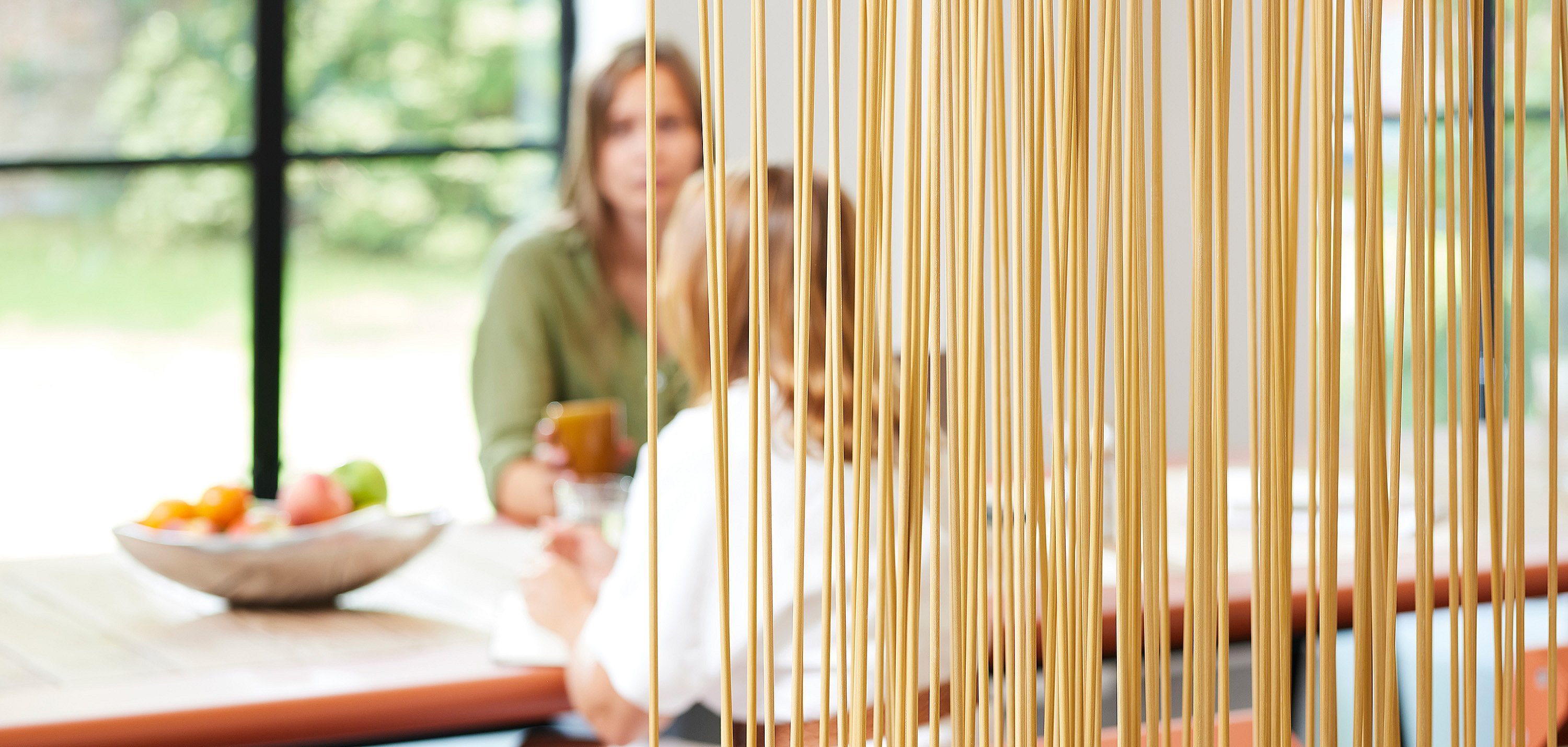 Close-up door een dicht scherm van slanke houtkleurige Sticks-kamerverdelers, met twee collega's die zitten en praten aan een tafel met een zachte focus erachter.