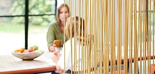 Close-up door een dicht scherm van slanke houtkleurige Sticks-kamerverdelers, met twee collega's die zitten en praten aan een tafel met een zachte focus erachter.