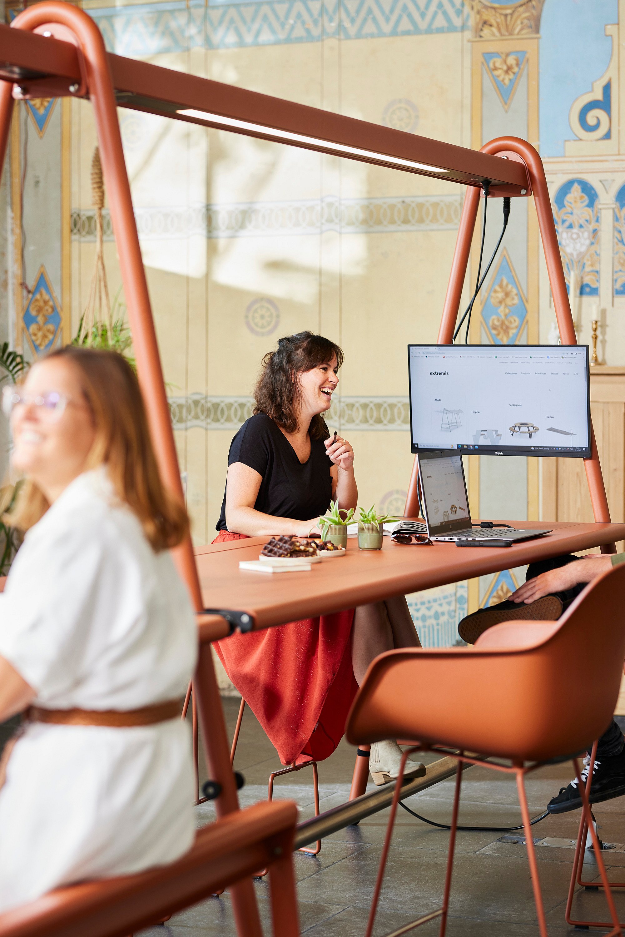 Vrouw lachend om een oranje AMAi tafel voorzien van een monitor en laptop, zittend tussen bijpassende stoelen in de kapel coworking space.