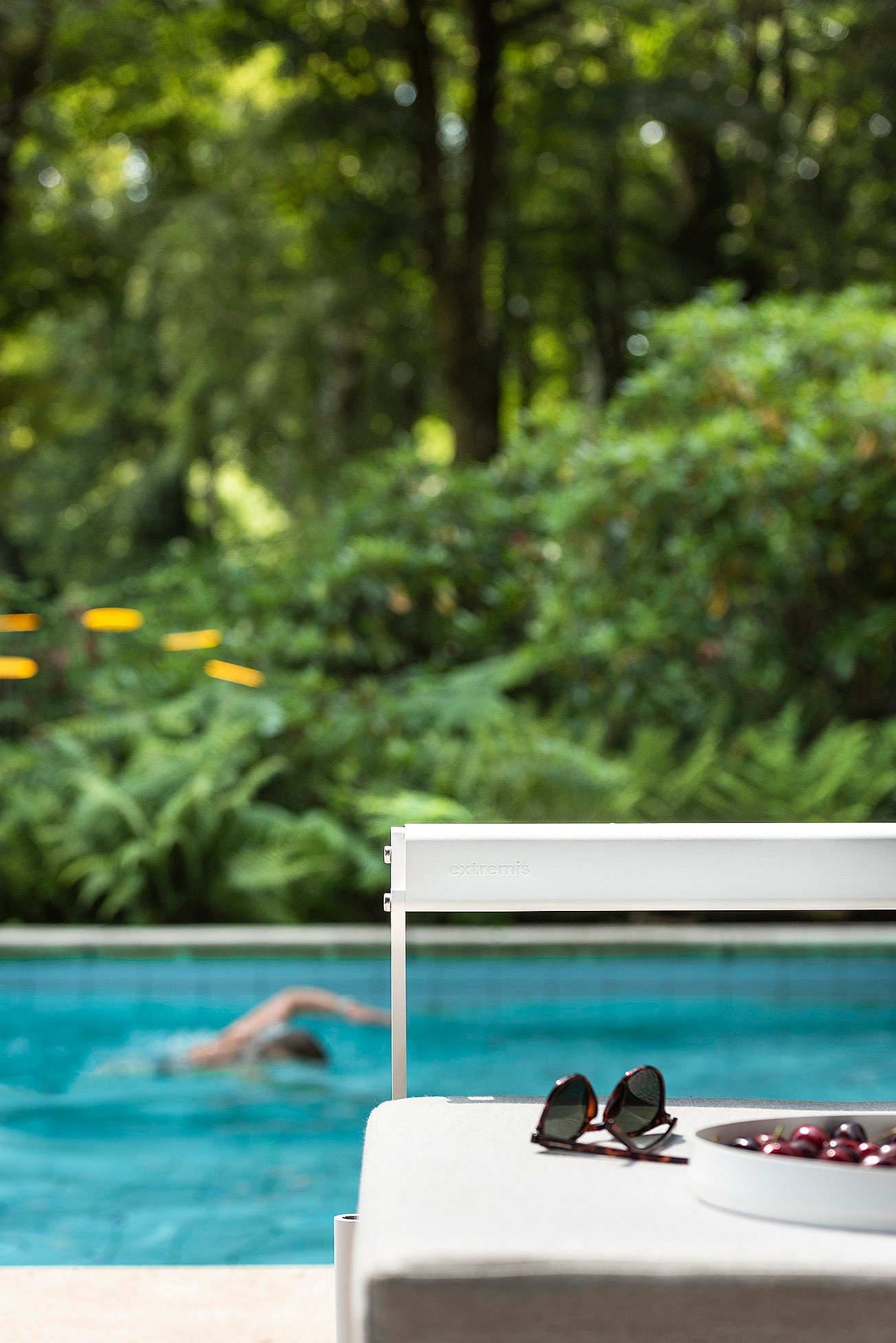 Gros plan du coin d’un sol extérieur blanc & luna avec des lunettes de soleil et un bol de cerises, tandis qu’un nageur se déplace dans la piscine turquoise sur le fond flou.