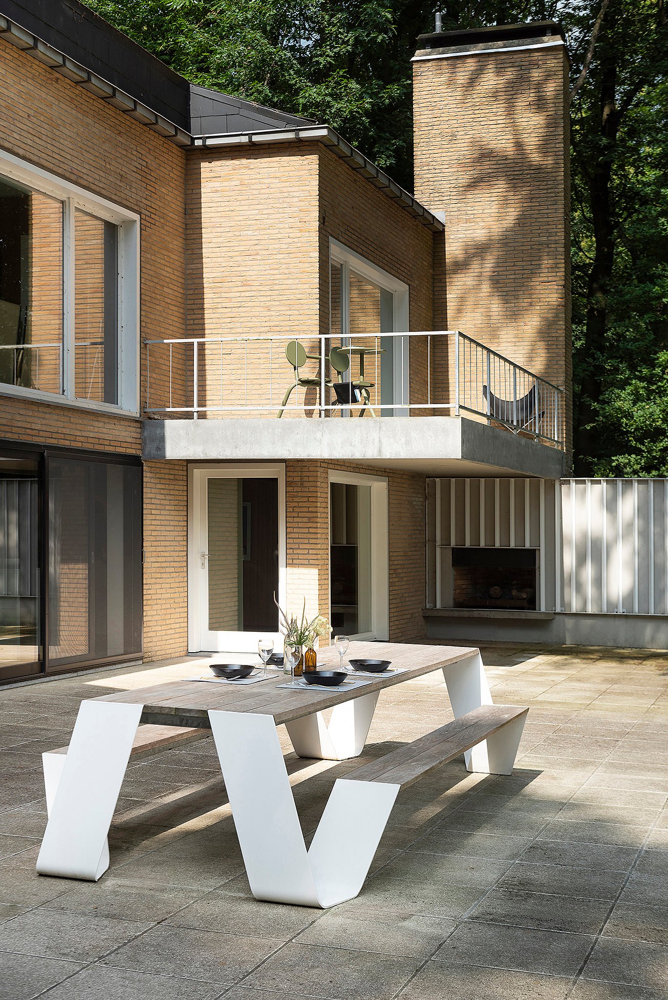 Large vue sur la terrasse de la villa avec une longue table de pique-nique Hopper au premier plan et un petit bistroo situé sur le balcon au-dessus, encadré par des murs en briques, de grandes fenêtres et des arbres environnants.