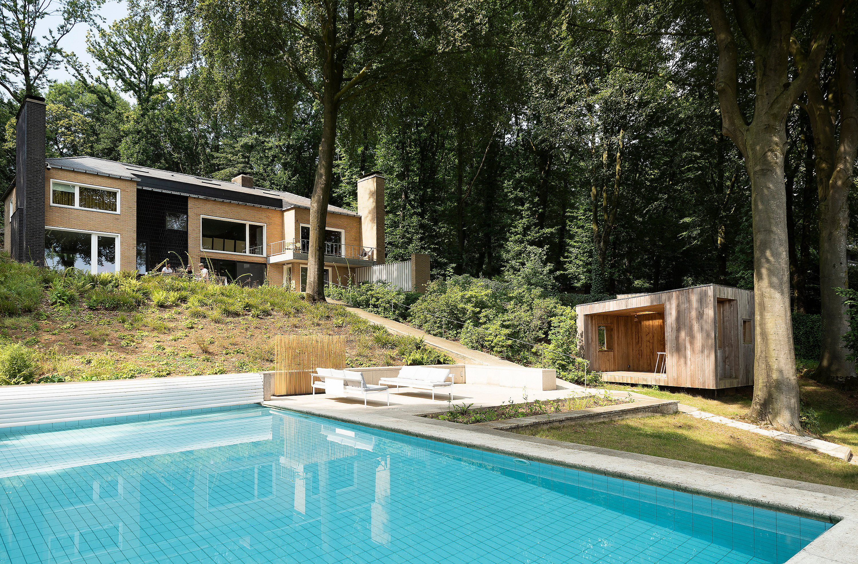 Villa moderniste en brique située dans une colline boisée au-dessus d’une grande piscine, avec des meubles de salon extérieurs blancs sur la terrasse inférieure et un petit pavillon de jardin en bois à droite.