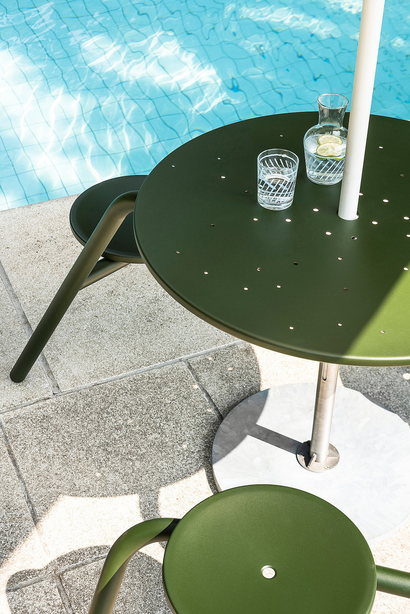 Close-up of a bottle-green round Virus table with matching stools beside the pool, topped with a glass carafe, drinking glass, and white acacia pole.