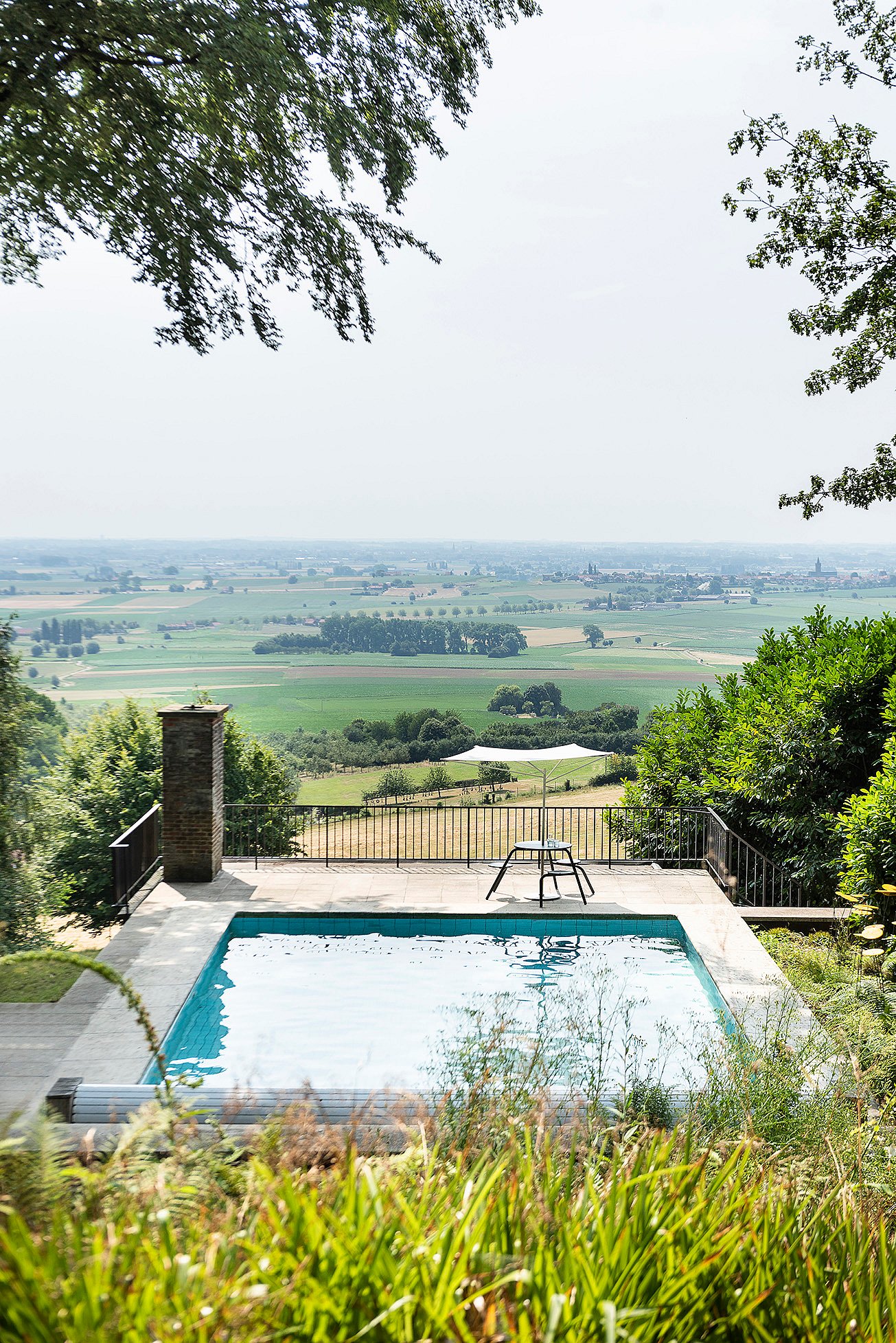 Piscine au sommet d’une colline et petite table ronde extérieure sur une terrasse donnant sur des champs en mosaïque et des villages lointains, encadrés par des arbres et des arbustes.