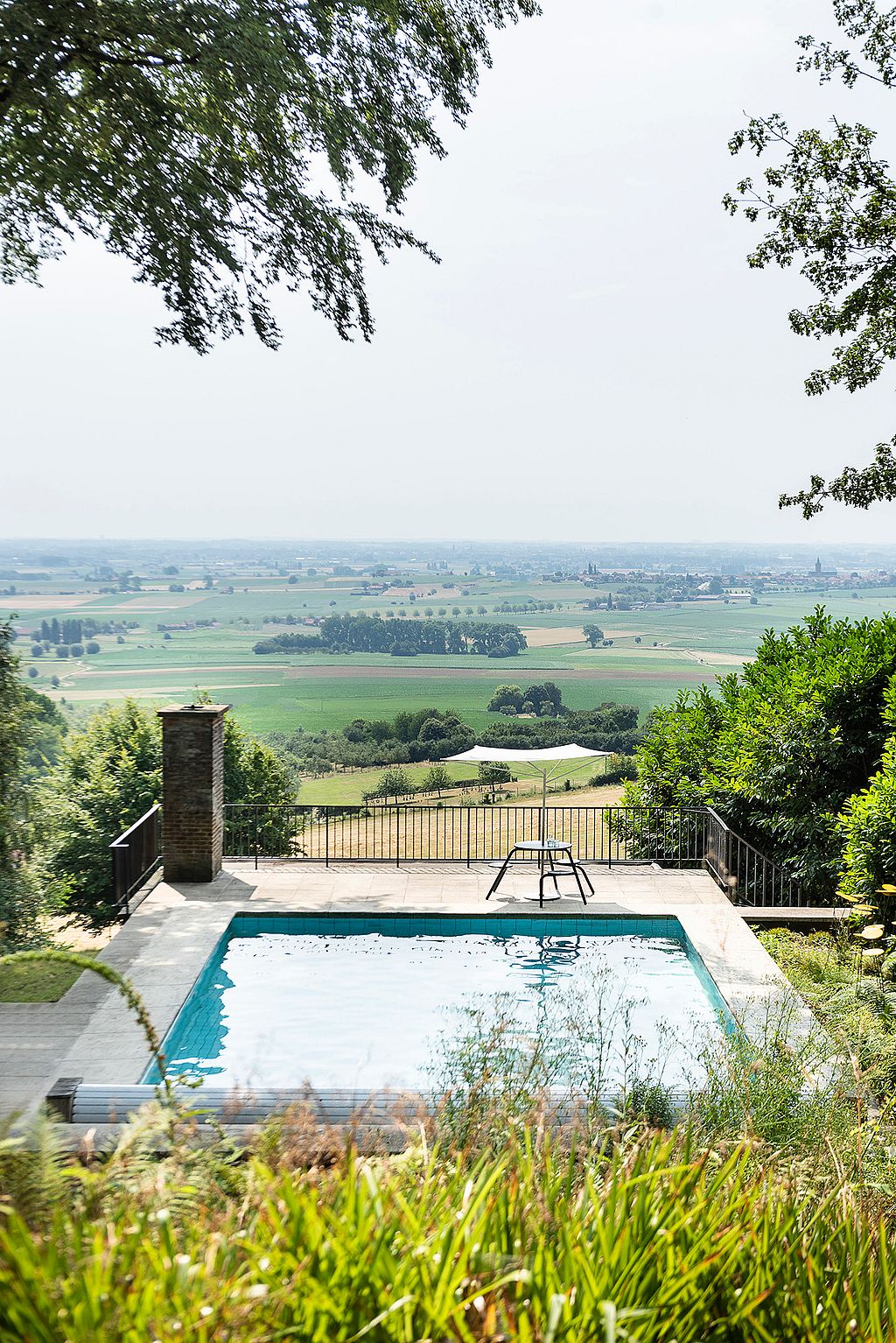 Piscine au sommet d’une colline et petite table ronde extérieure sur une terrasse donnant sur des champs en mosaïque et des villages lointains, encadrés par des arbres et des arbustes.