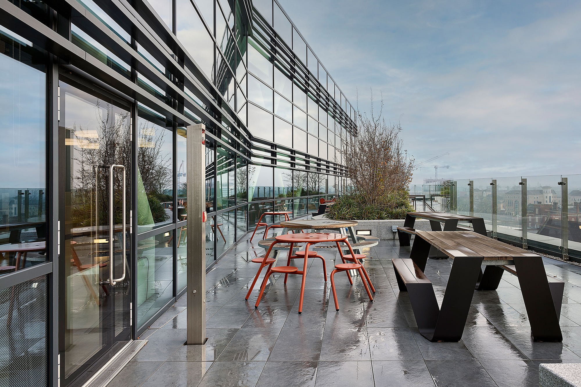 Terrasse de bureau humide sur le toit à côté d’une façade en verre courbée, meublée d’une table de pique-nique Hopper noire et de tables rondes extérieures rouges avec des tabourets attachés, ainsi que des jardinières et une balustrade en verre donnant sur la ville.