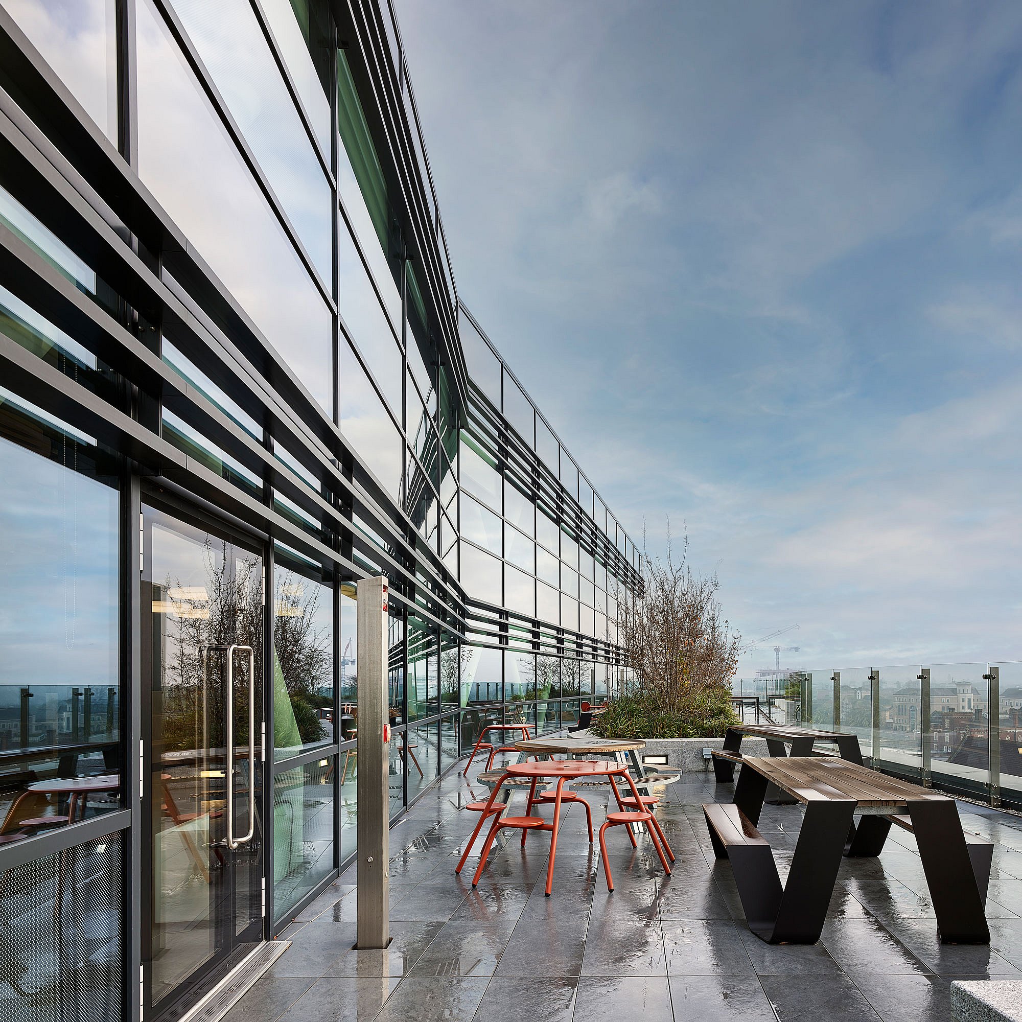 Weitblick einer Dachterrasse vor einem modernen Bürogebäude aus Glas, mit einem schwarzen Picknicktisch, roten runden Hockern und Tischsets, großen Töpfen und einem vom Regen unter einem hellblauen Himmel verdunkelten Pflaster.