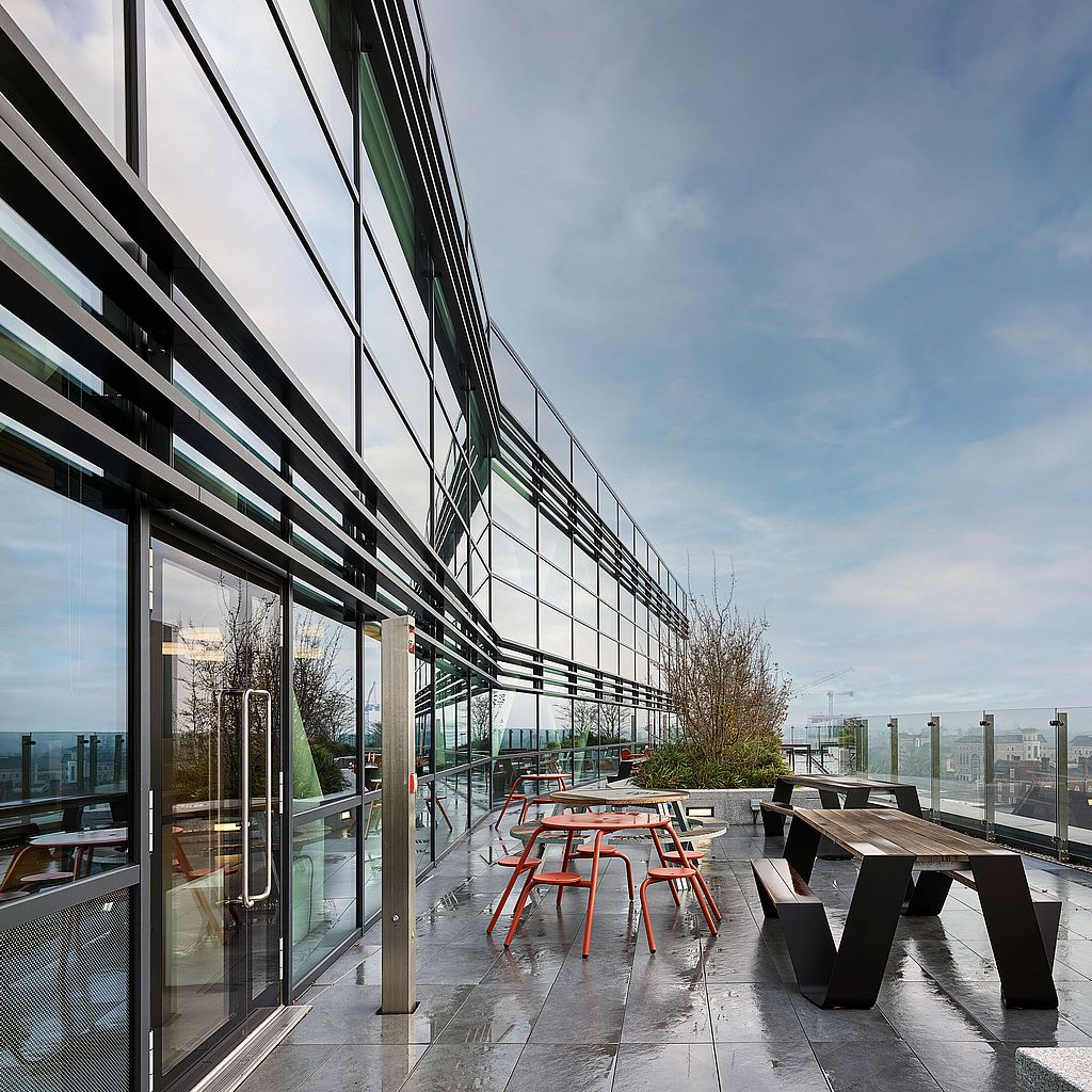 Weitblick einer Dachterrasse vor einem modernen Bürogebäude aus Glas, mit einem schwarzen Picknicktisch, roten runden Hockern und Tischsets, großen Töpfen und einem vom Regen unter einem hellblauen Himmel verdunkelten Pflaster.
