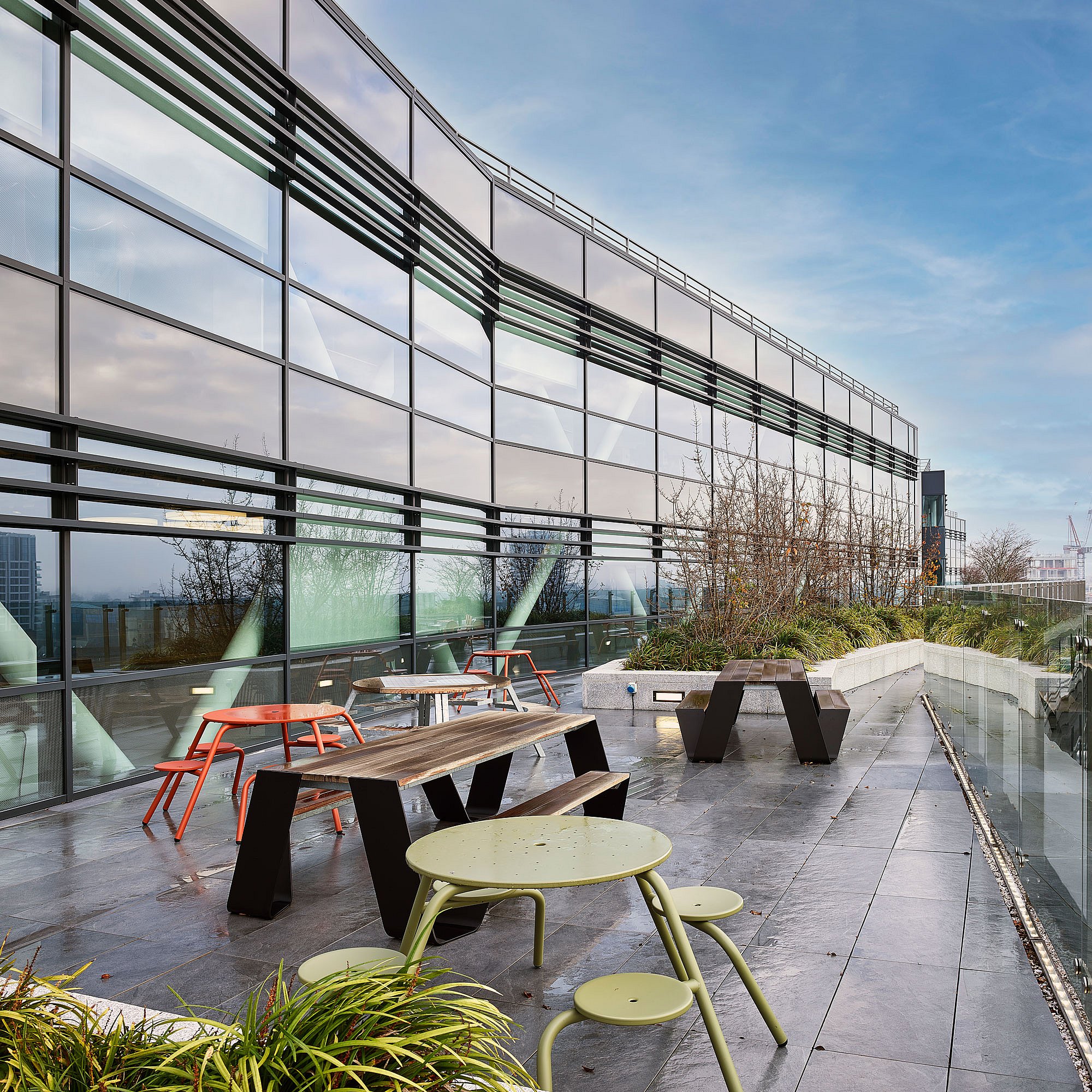 Große Dachterrasse mit zwei schwarzen Hopper-Picknicktischen, einem hellgrünen runden Tisch mit im Vordergrund befestigten Hockern, roten runden Sitzen in der Nähe der Fassade und eingepflanzten Bordsteinen entlang des Gebäuderandes.