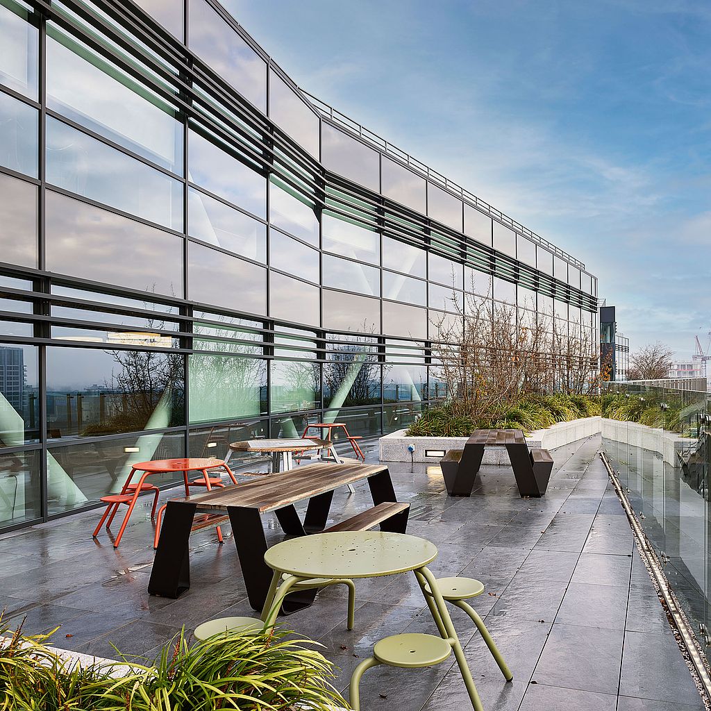 Große Dachterrasse mit zwei schwarzen Hopper-Picknicktischen, einem hellgrünen runden Tisch mit im Vordergrund befestigten Hockern, roten runden Sitzen in der Nähe der Fassade und eingepflanzten Bordsteinen entlang des Gebäuderandes.