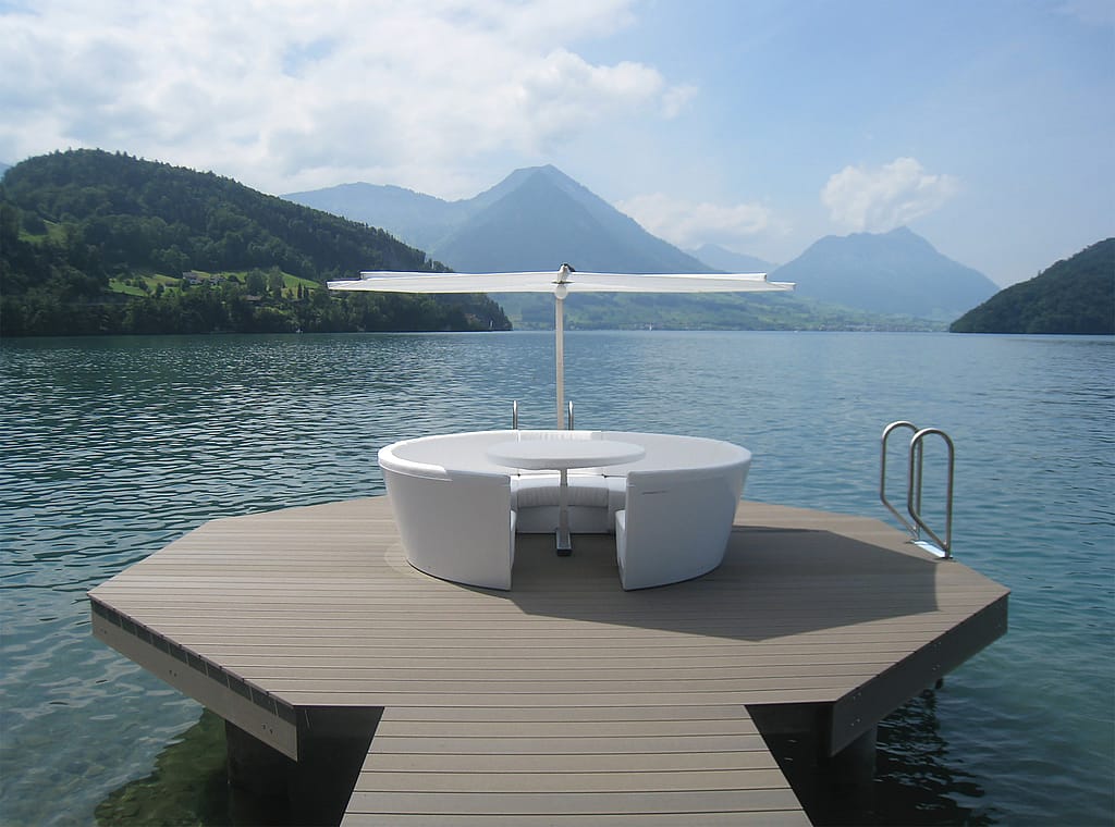 Vooraanzicht van een wit Kosmos ziteiland met ronde tafel en parasol op een terras aan het meer, omlijst door blauw water en gelaagde bergtoppen onder een heldere hemel.