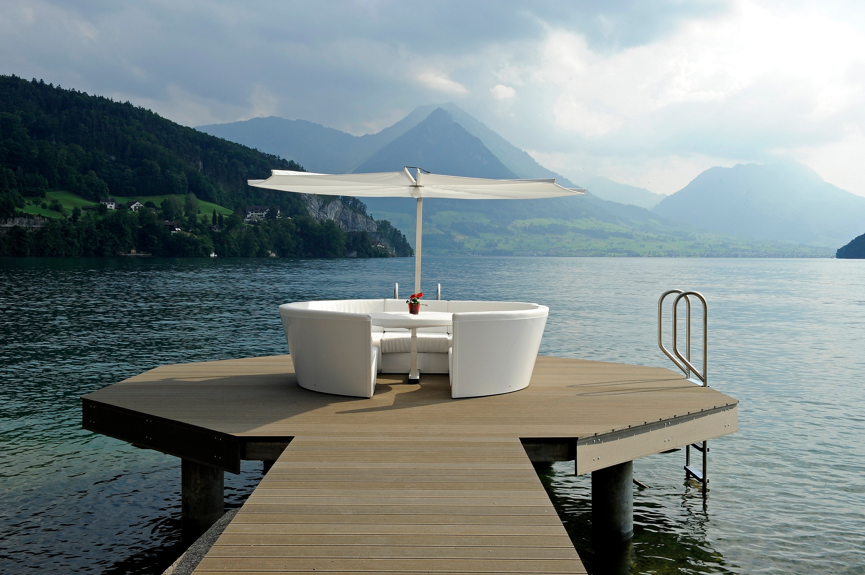 Salon de jardin Kosmos blanc avec table centrale et parasol, installé sur une petite plateforme en bois au bord d'un lac, entouré d'eaux calmes, avec des montagnes boisées et un ciel nuageux en arrière-plan.