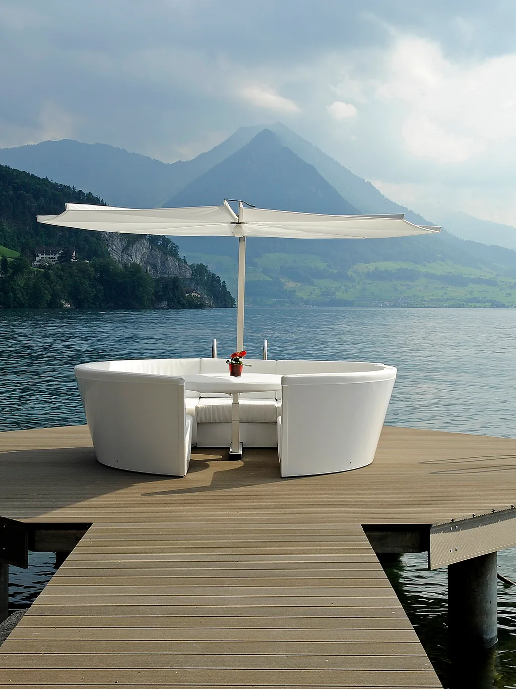 Salon de jardin Kosmos blanc avec table centrale et parasol, installé sur une petite plateforme en bois au bord d'un lac, entouré d'eaux calmes, avec des montagnes boisées et un ciel nuageux en arrière-plan.