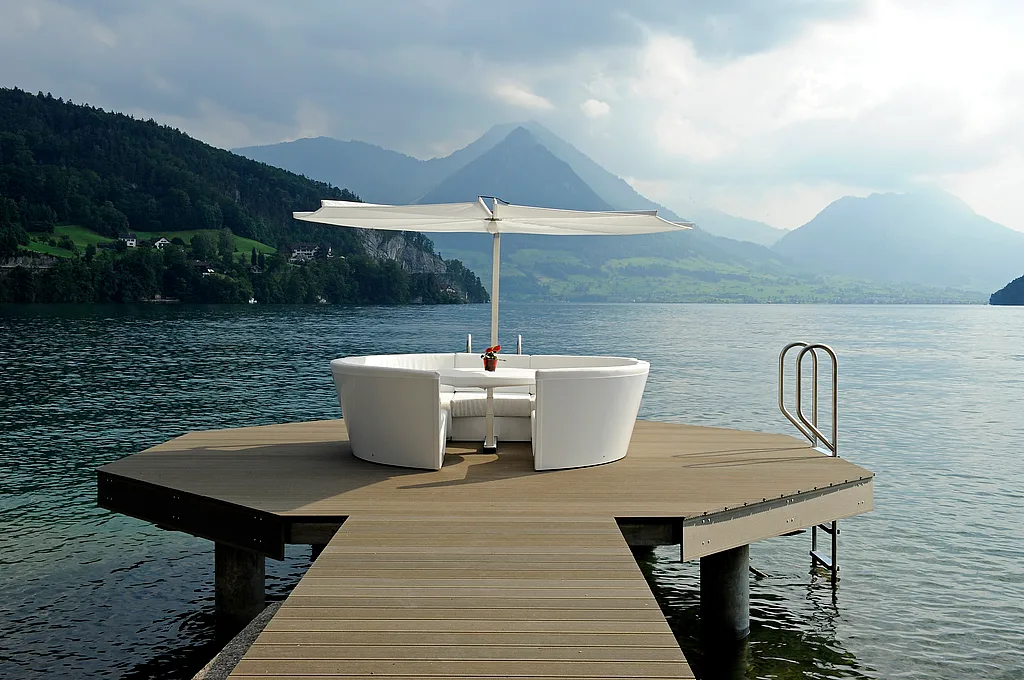 Witte Kosmos loungeset met centrale tafel en parasol op een klein houten platform aan het meer, omgeven door kalm water met beboste bergen en een bewolkte hemel op de achtergrond.