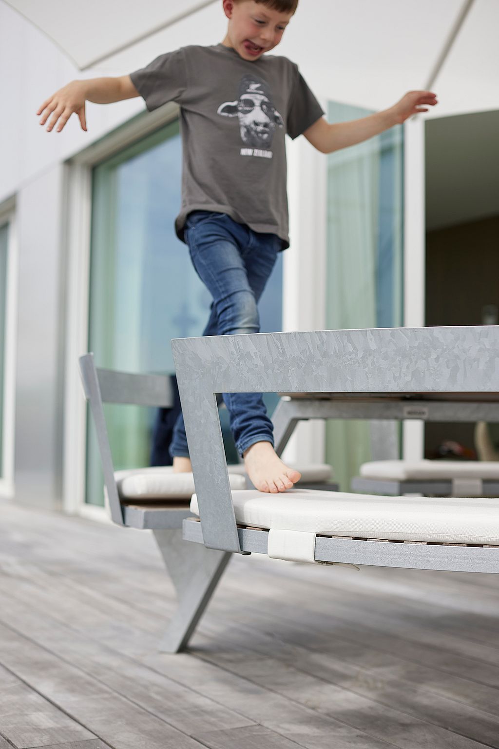 Kind balanceert op blote voeten op de kussenbank van een ronde buitentafel naast de glazen deuren van het penthouse-terras.