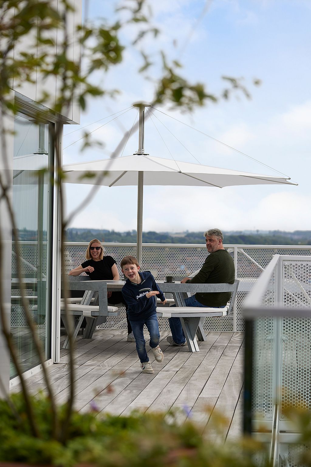Jonge jongen die op blote voeten naar de camera rent terwijl twee volwassenen aan een ronde Gargantua-tafel zitten onder een grote witte parasol op het dakterras.