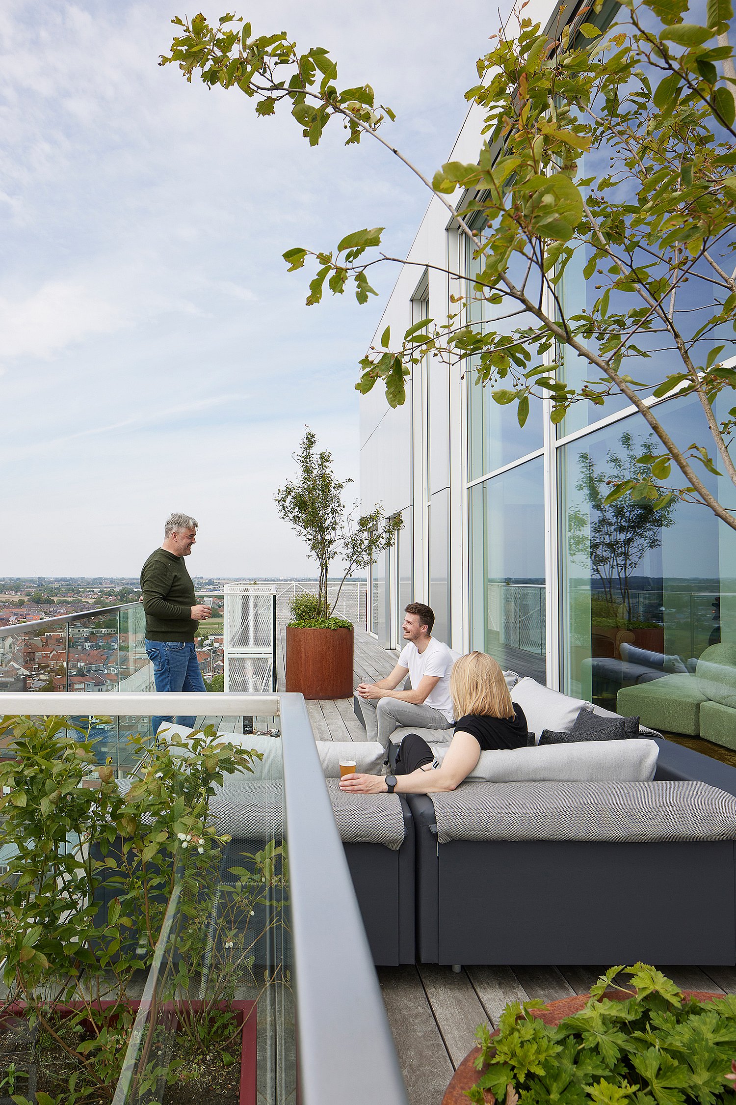 Drie mensen ontspannen op een modulaire grijze Walrus-buitenbank op een dakterras naast een glazen gevel, met plantenbakken, bomen en een panoramisch uitzicht over de stad om hen heen.