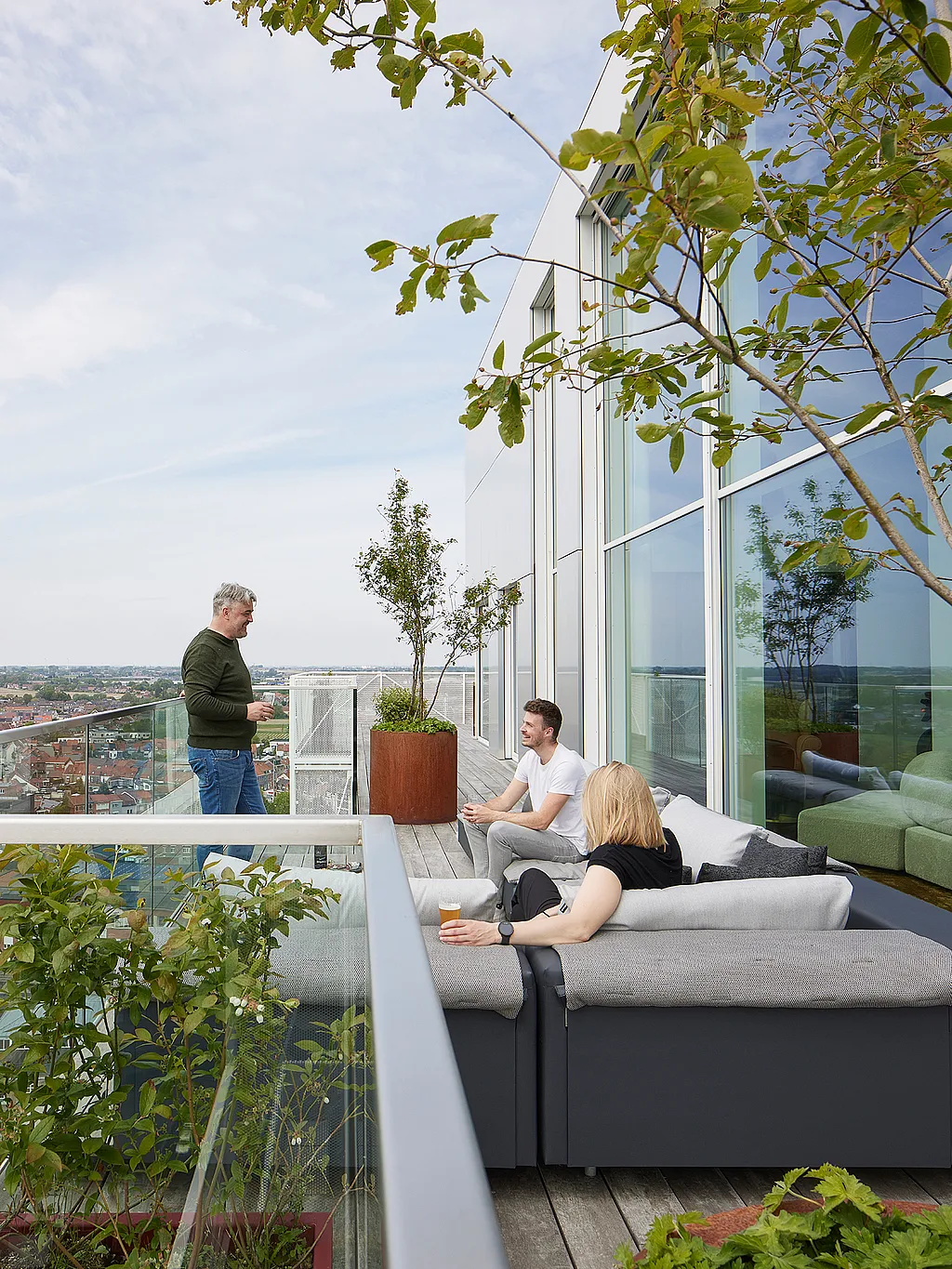 Drie mensen ontspannen op een modulaire grijze Walrus-buitenbank op een dakterras naast een glazen gevel, met plantenbakken, bomen en een panoramisch uitzicht over de stad om hen heen.
