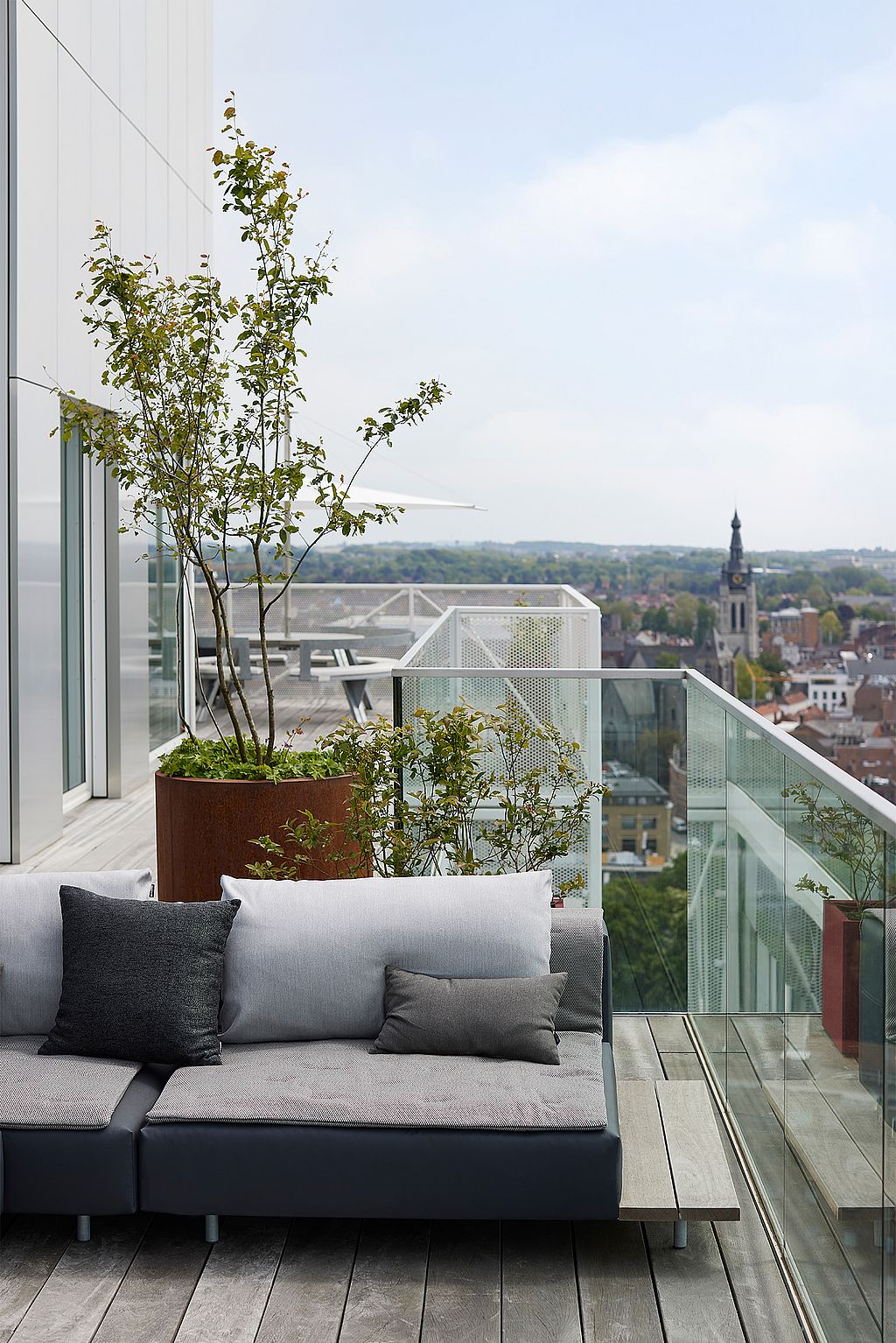 Hoek van een grijze walrus met kussens op een dakterras, omlijst door roestkleurige plantenbakken en een glazen balustrade met uitzicht op de stad en de kerktoren daarachter.