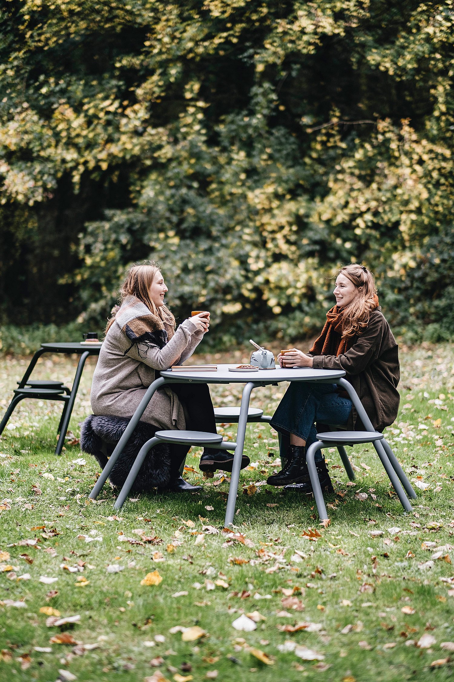   Zwei Frauen in Mänteln und Schals sitzen an einem Virus-Tisch, trinken heiße Getränke und unterhalten sich auf einer Wiese, umgeben von Herbstbäumen.
