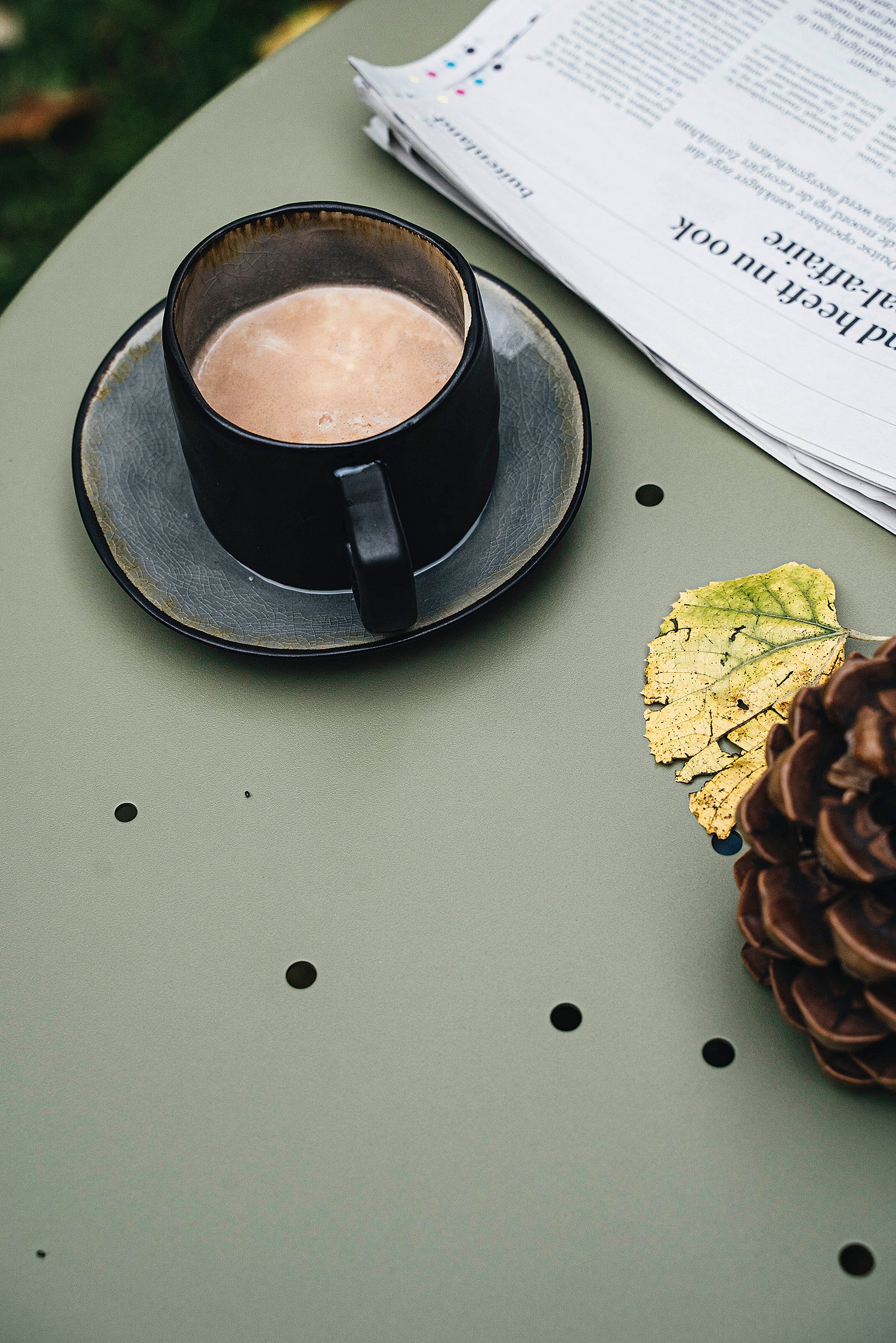   Eine schwarze Tasse heißer Schokolade auf einer Untertasse neben einer gefalteten Zeitung, einem gelben Blatt und einem Tannenzapfen auf einem olivgrünen Gartentisch.