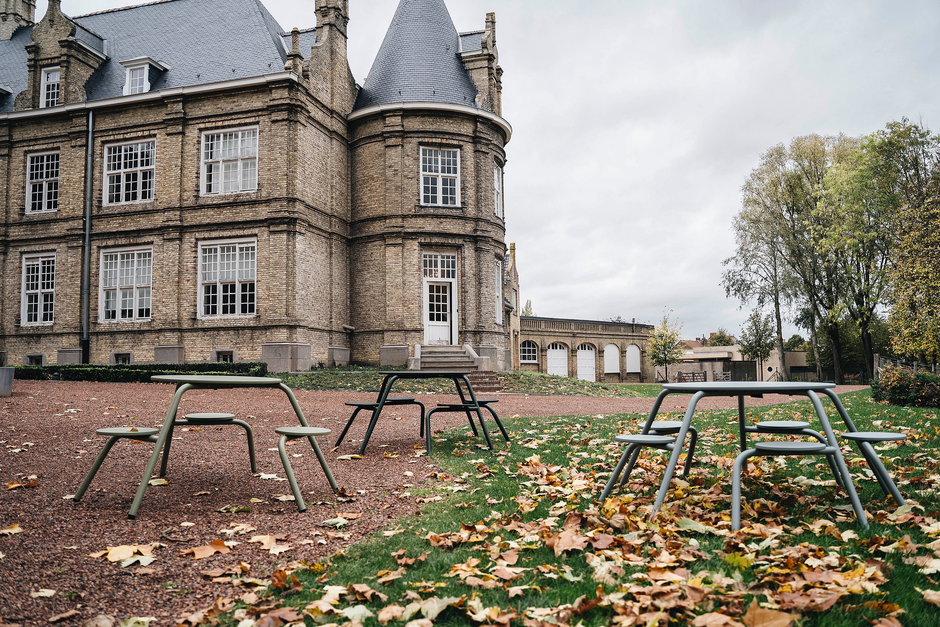Historic stone castle the Warande with turrets and tall windows, seen behind several green and black Virus picnic tables scattered across a gravel path and leaf-covered lawn on an overcast autumn day