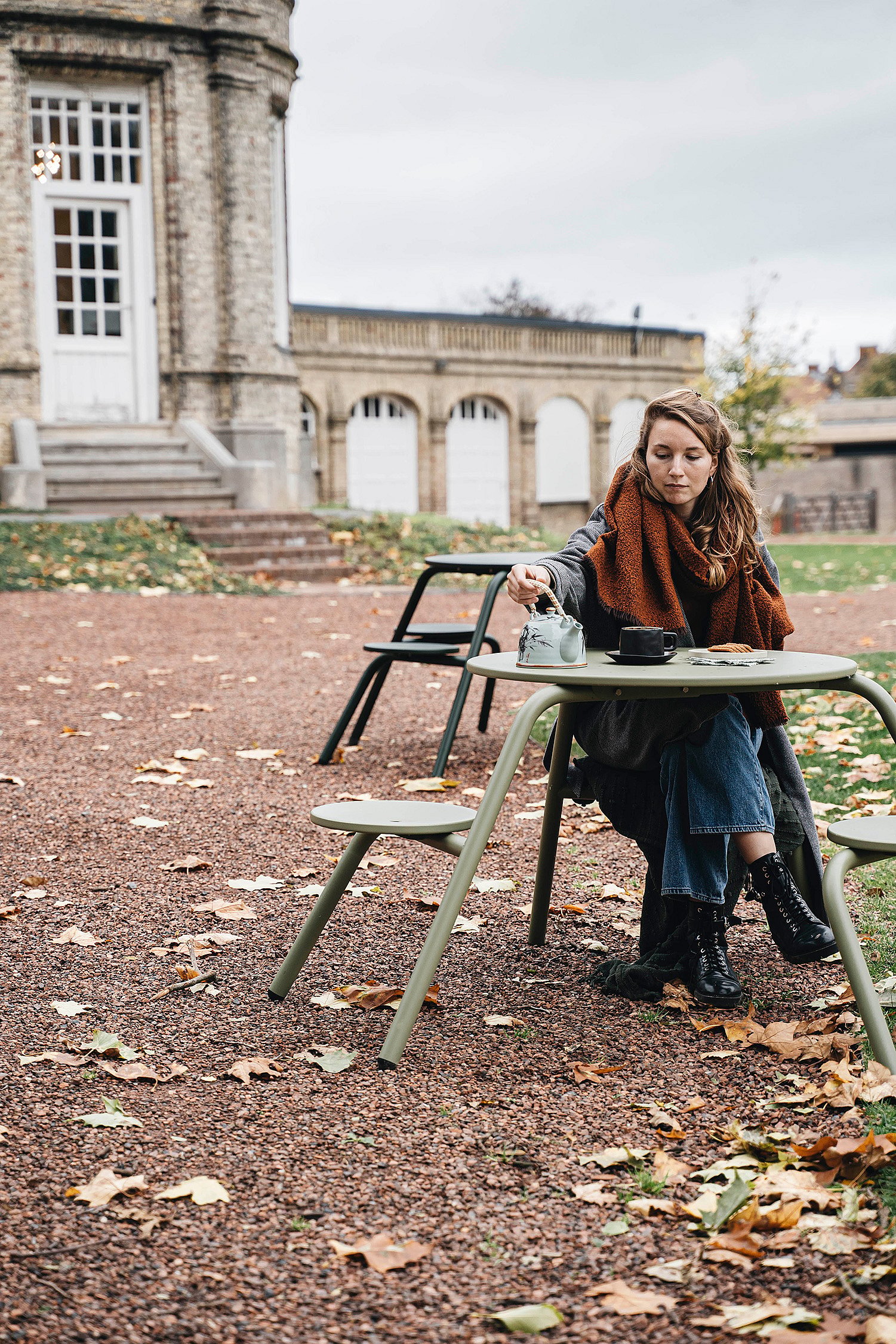   Eine Frau mit rostfarbenem Schal sitzt allein an einem grünen Virus-Picknicktisch in einem mit Laub bedeckten Kieshof und schenkt sich Tee vor dem Schloss ein.