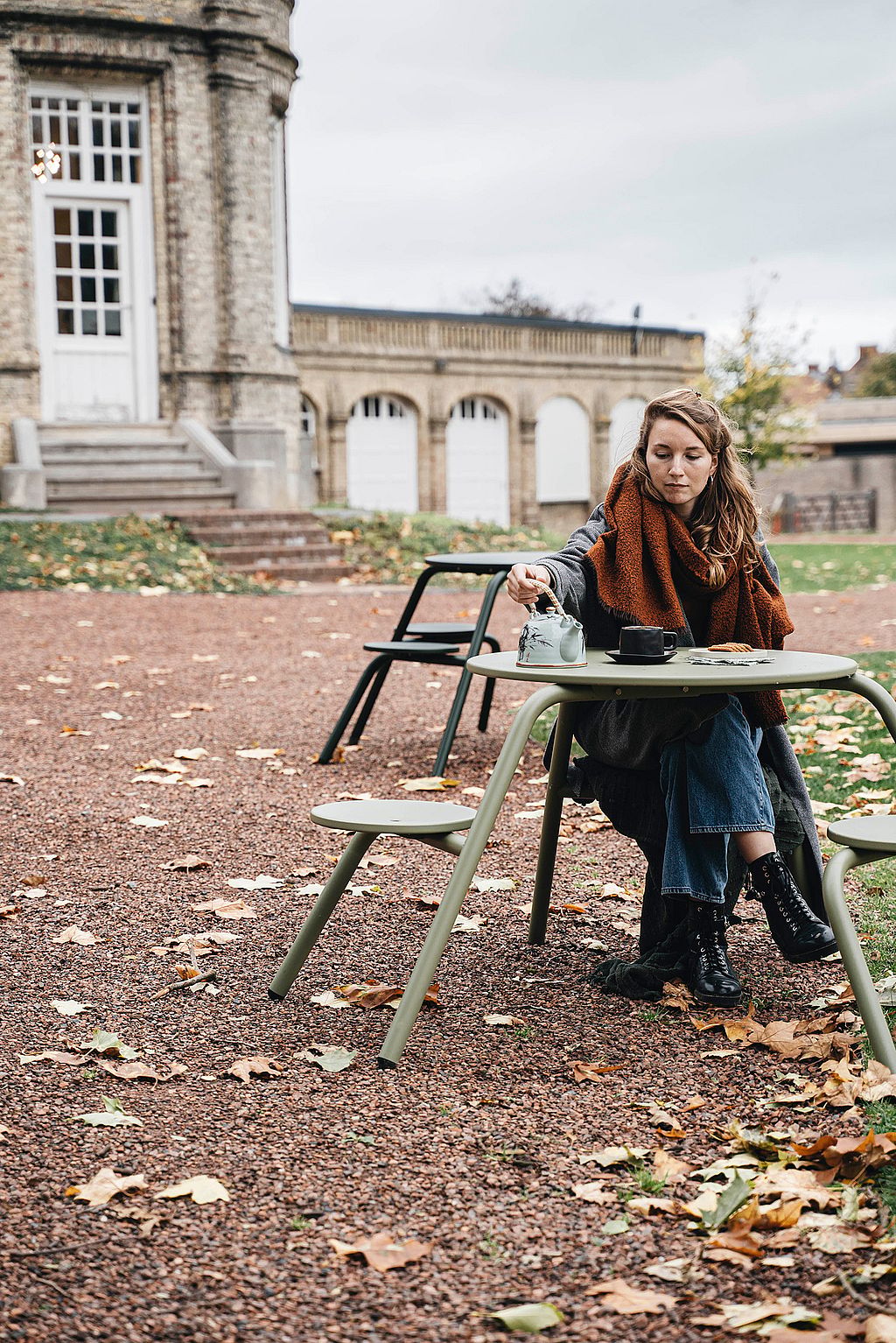 Eine Frau mit rostfarbenem Schal sitzt allein an einem grünen Virus-Picknicktisch in einem mit Laub bedeckten Kieshof und schenkt sich Tee vor dem Schloss ein.