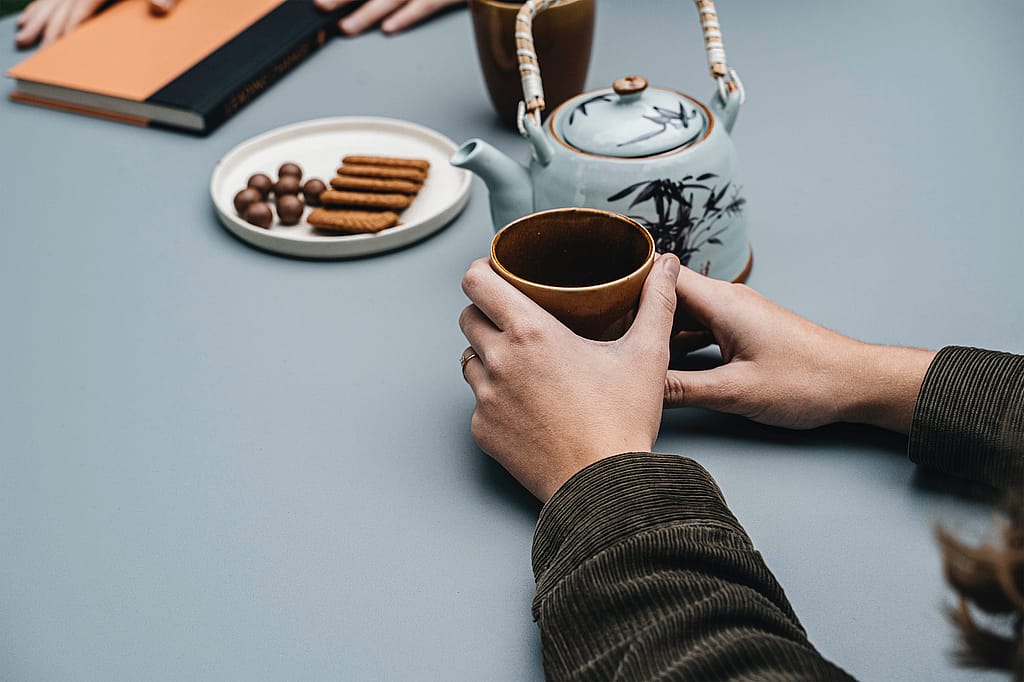 Nahaufnahme von Händen, die eine braune Tasse halten, neben einer Keramik-Teekanne, Keksen, Pralinen und einem Notizbuch auf einer glatten, grauen Tischplatte.