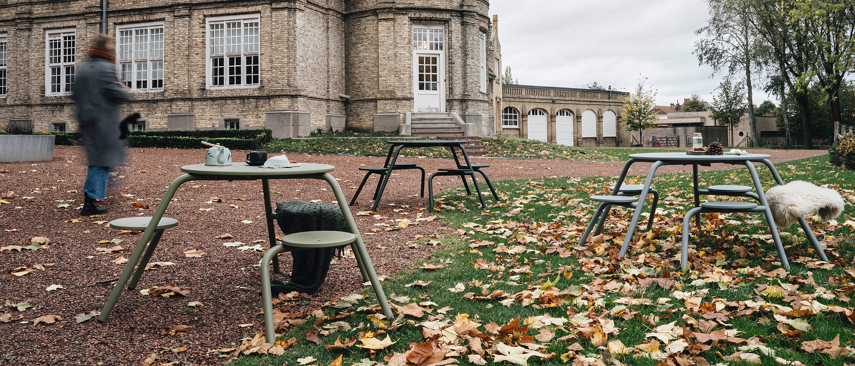   Weite Herbstszene: Mehrere Virus-Picknicktische sind auf Kies und Gras vor dem Schloss verteilt. Gefallenes Laub bedeckt den Boden, und eine verschwommene Person geht vorbei.