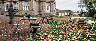 Weite Herbstszene: Mehrere Virus-Picknicktische sind auf Kies und Gras vor dem Schloss verteilt. Gefallenes Laub bedeckt den Boden, und eine verschwommene Person geht vorbei.