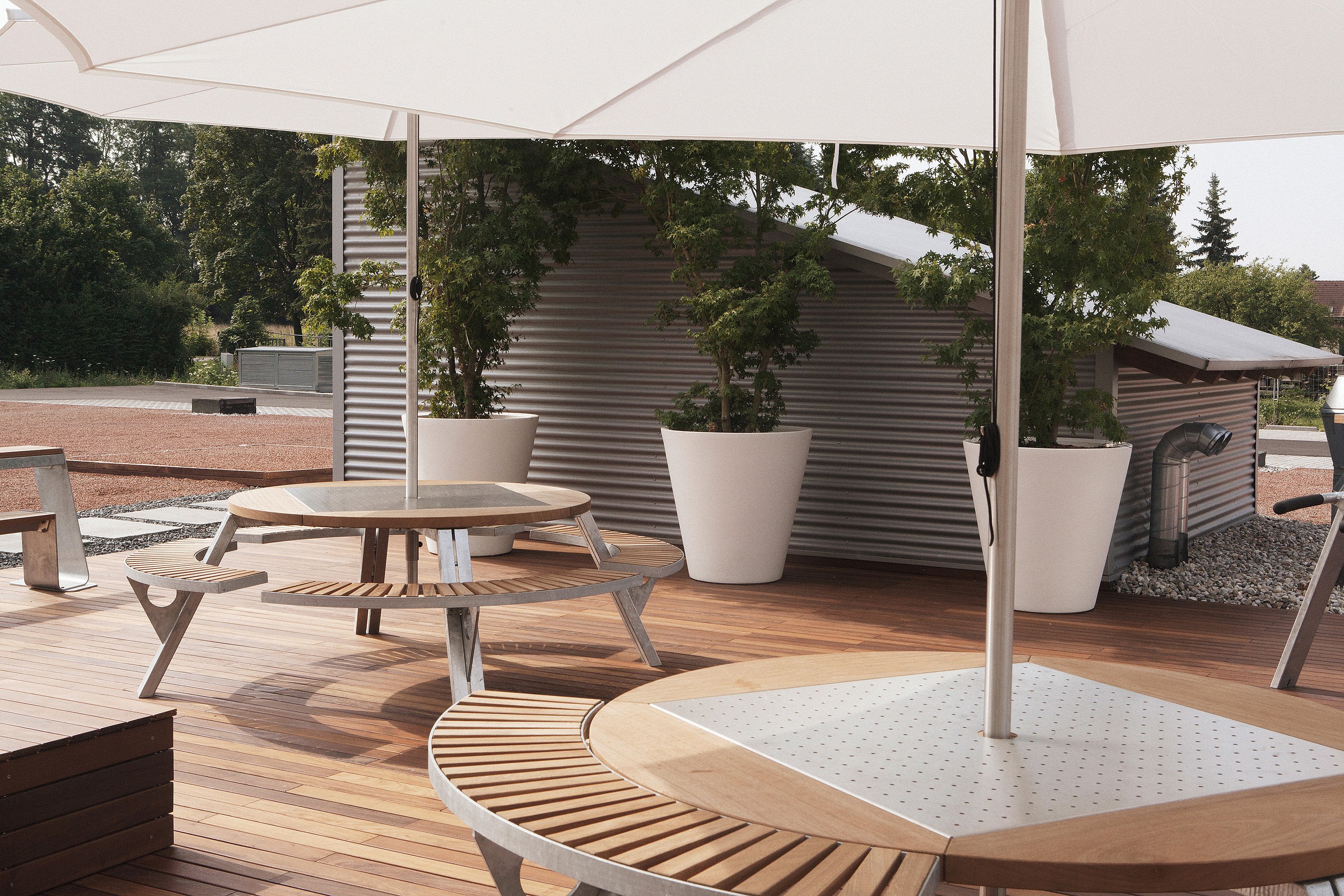 Close view of gargantua tables and attached benches on a timber deck beneath white inumbrinas, with tall white planters and a corrugated garden structure behind.