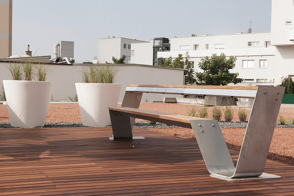 Close-up of a long hopper with galvanized steel legs on a sunlit rooftop terrace, framed by large white planters and low gravel planting beds.