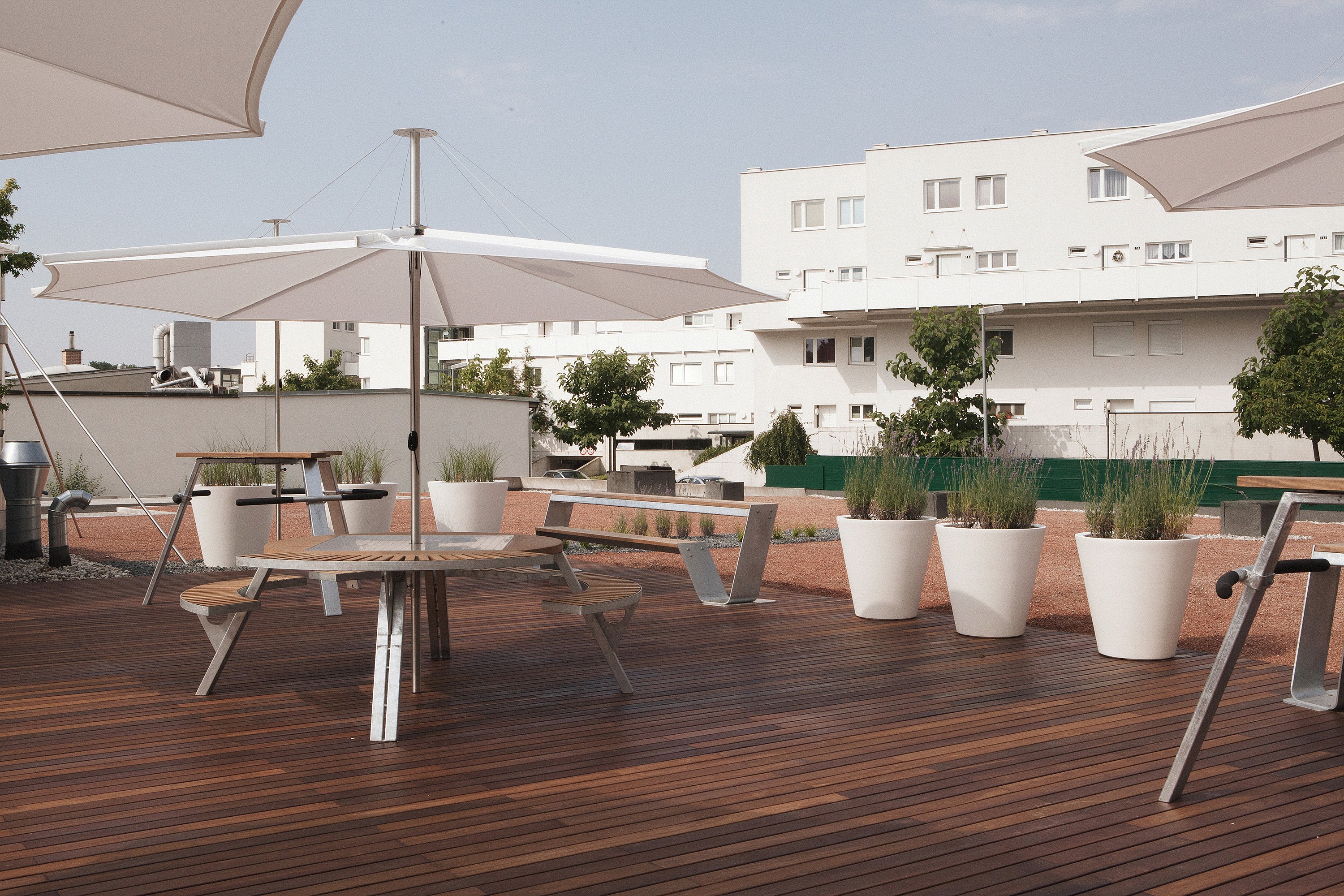 Round outdoor gargantua table with attached benches beneath a wide white parasol on a rooftop deck, with large white plant pots, long benches, and white residential buildings in the background.