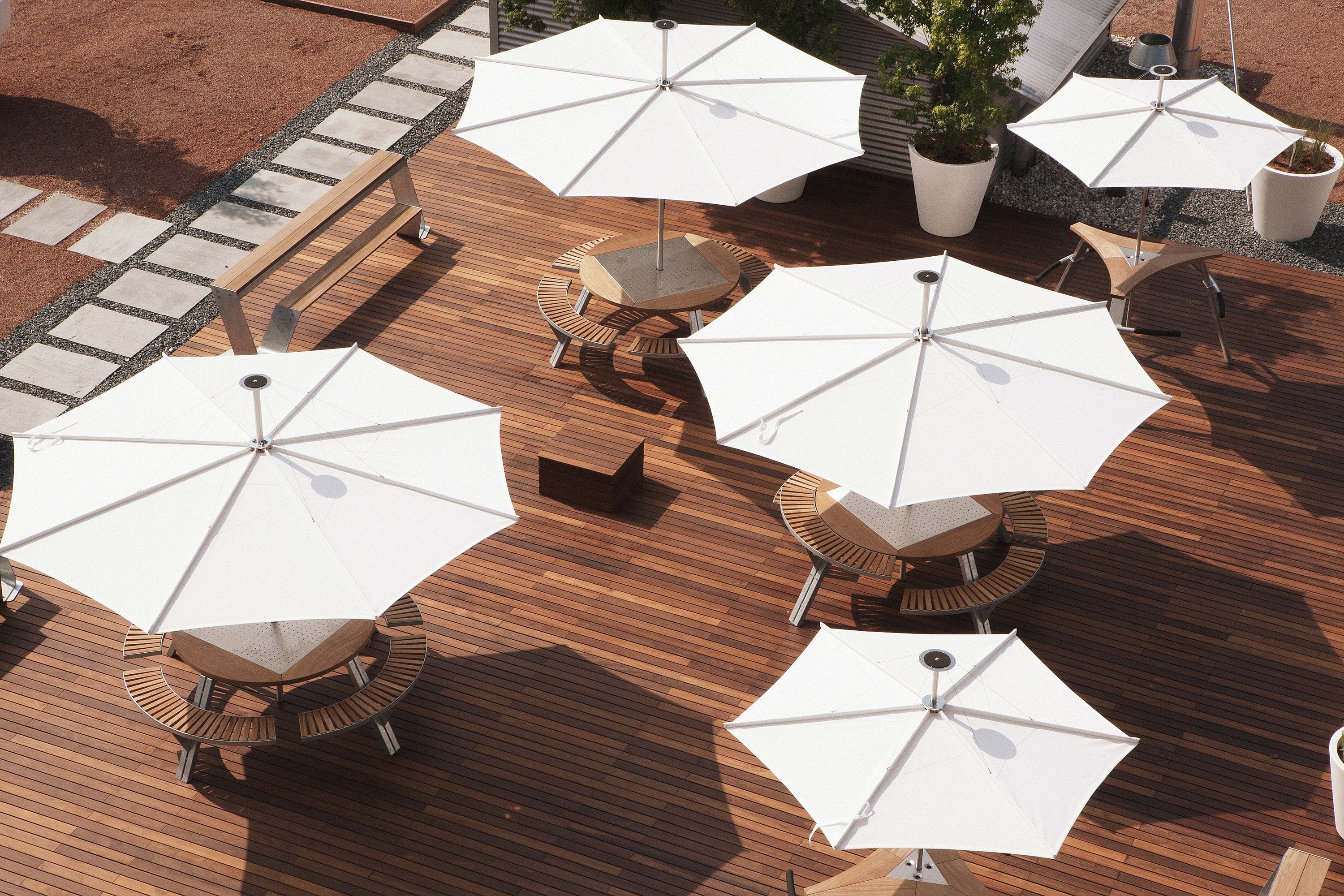 Draufsicht auf eine große hölzerne Dachterrasse mit vier weißen Sonnenschirmen, die runde Picknicktische beschatten, neben einer langen Bank, übergroßen weißen Pflanzgefäßen und einem abgestuften Gehweg durch roten Kies.