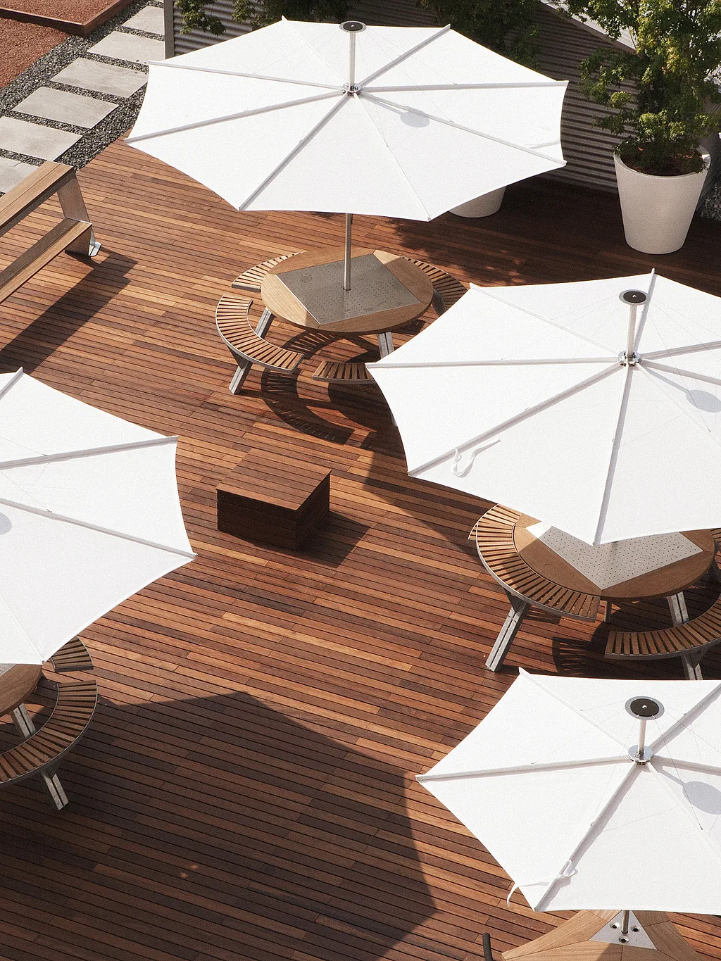Draufsicht auf eine große hölzerne Dachterrasse mit vier weißen Sonnenschirmen, die runde Picknicktische beschatten, neben einer langen Bank, übergroßen weißen Pflanzgefäßen und einem abgestuften Gehweg durch roten Kies.