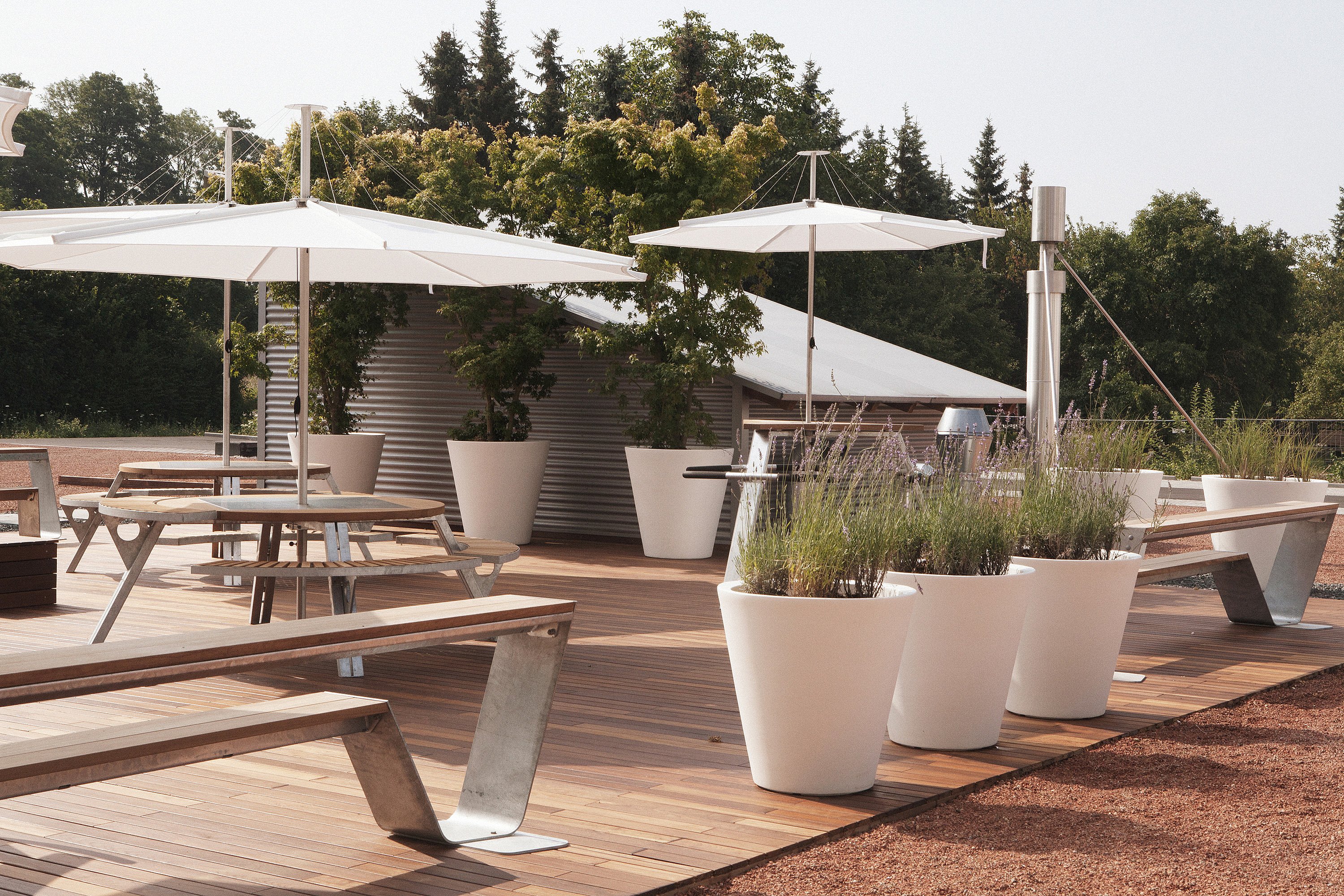 Wide view of the social terrace with gargantua tables, white inumbrinas, hoppers, and oversized white plant pots, bordered by red gravel and trees