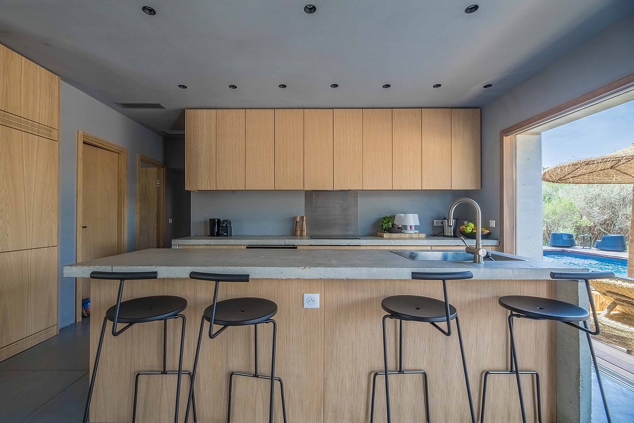   Een minimalistische keuken met licht houten kasten en een betonnen kookeiland met vier zwarte Tiki barkrukken, recht van voren gezien met het zwembadterras zichtbaar door het open zijraam.
