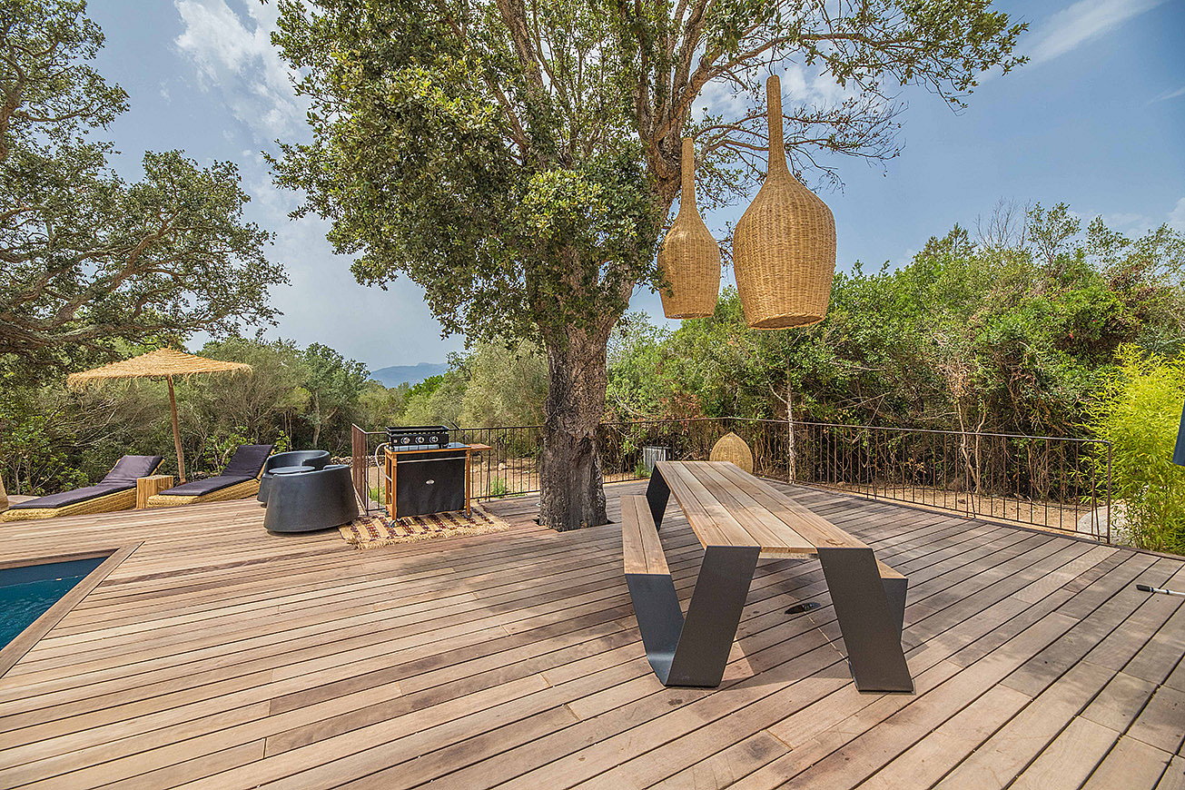  Een lange houten Hopper picknicktafel op een ruim houten terras onder een volwassen boom, met geweven hanglampen erboven, ligstoelen en een parasol naast het zwembad, en weelderig groen eromheen.