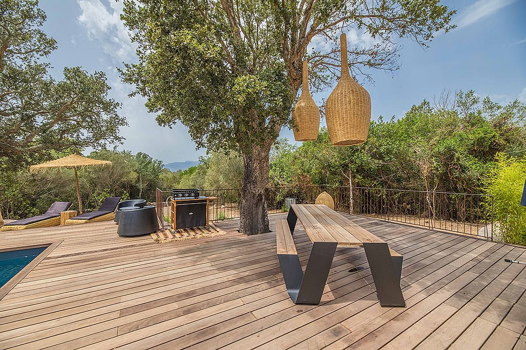 Een lange houten Hopper picknicktafel op een ruim houten terras onder een volwassen boom, met geweven hanglampen erboven, ligstoelen en een parasol naast het zwembad, en weelderig groen eromheen.