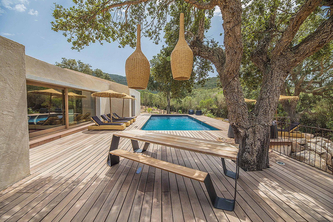 Villa moderne en béton avec piscine rectangulaire et large terrasse en bois. Au premier plan, une table de pique-nique Hopper en bois est placée sous un grand arbre, éclairée par deux suspensions en osier.
