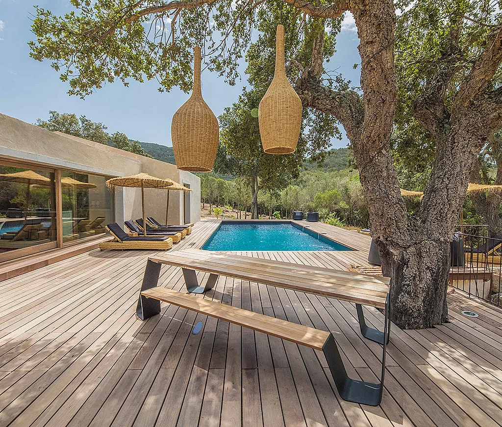 Villa moderne en béton avec piscine rectangulaire et large terrasse en bois. Au premier plan, une table de pique-nique Hopper en bois est placée sous un grand arbre, éclairée par deux suspensions en osier.